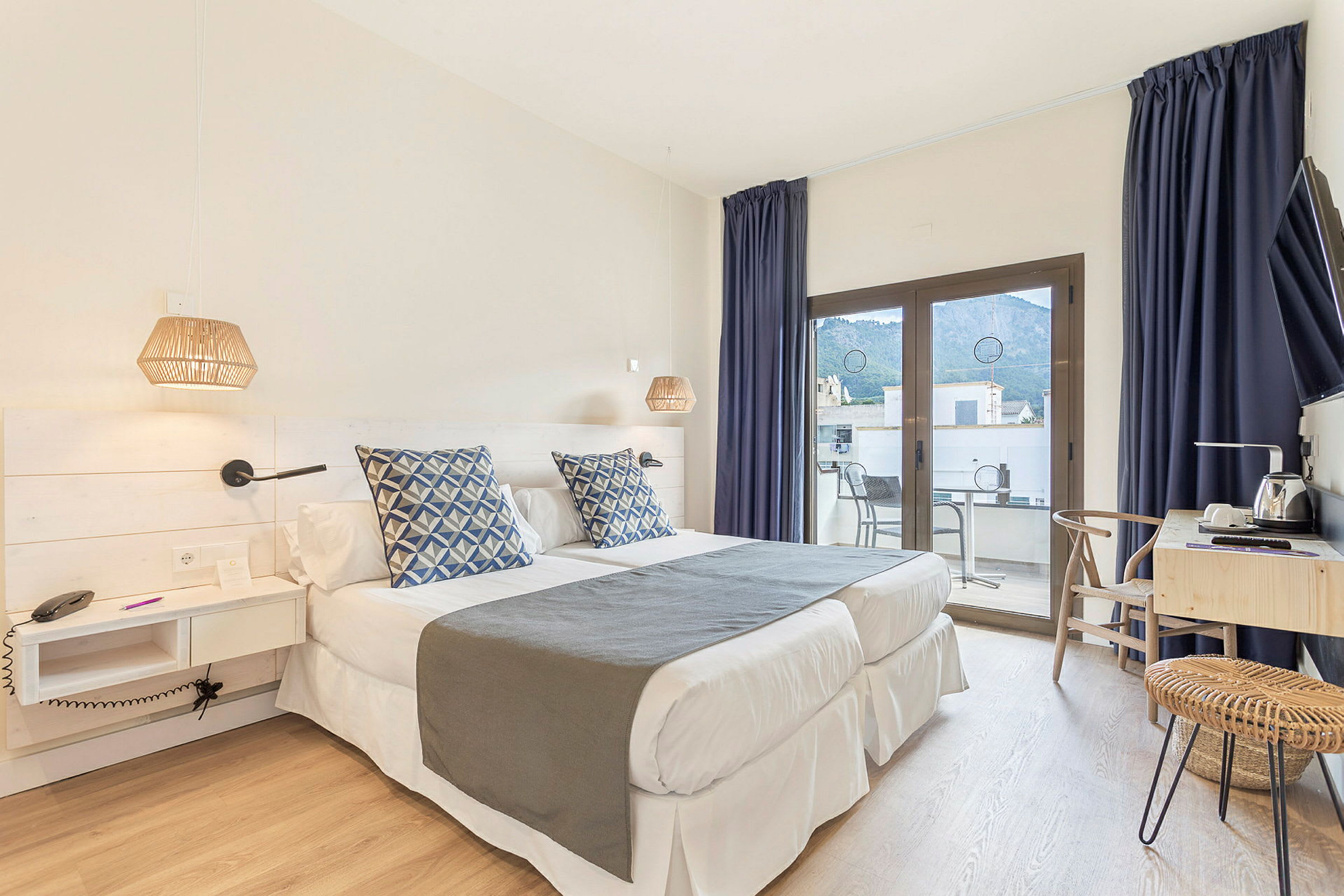 Et hotelværelse med to enkeltsenge, blå dekorationspuder, skrivebord og en altan med udsigt. (AI-genereret tekst)