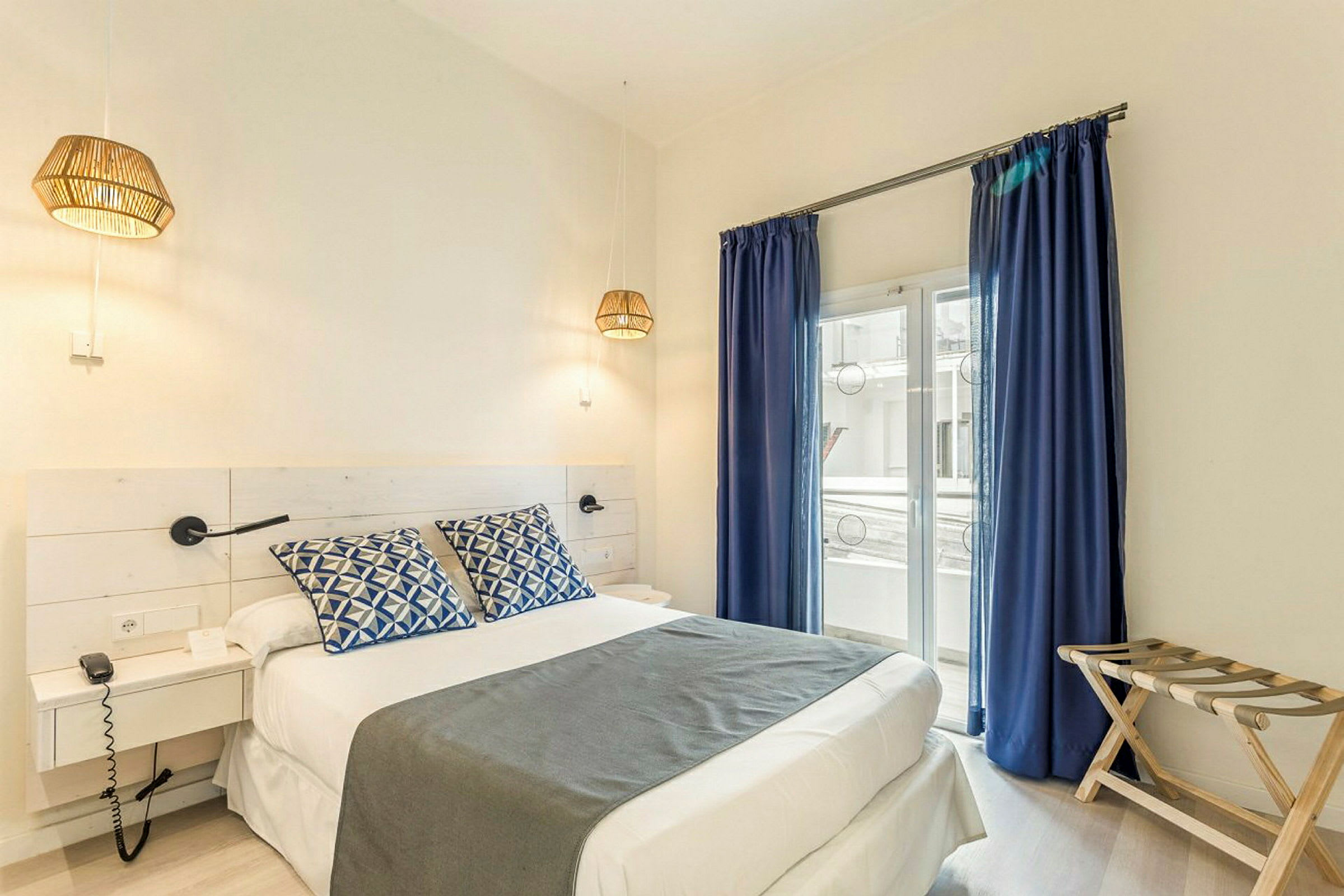Et hotelværelse med en dobbeltseng, blå gardiner og vindue mod en balkon. (AI-genereret tekst)