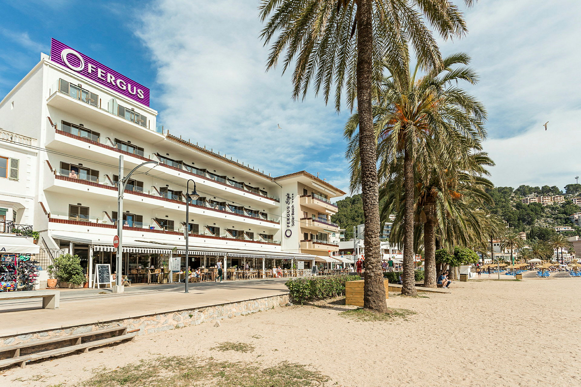 Et hotel med flere etager og altaner ligger ved en sandstrand, omgivet af palmer. (AI-genereret tekst)