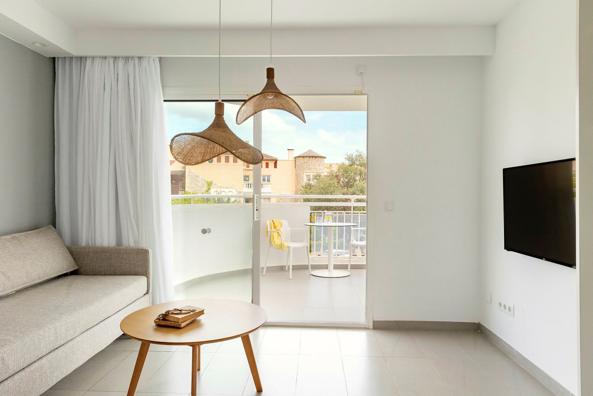 En stue med en sofa, et rundt bord og to hængende lamper samt udsigt til en balkon med møbler. (AI-genereret tekst)