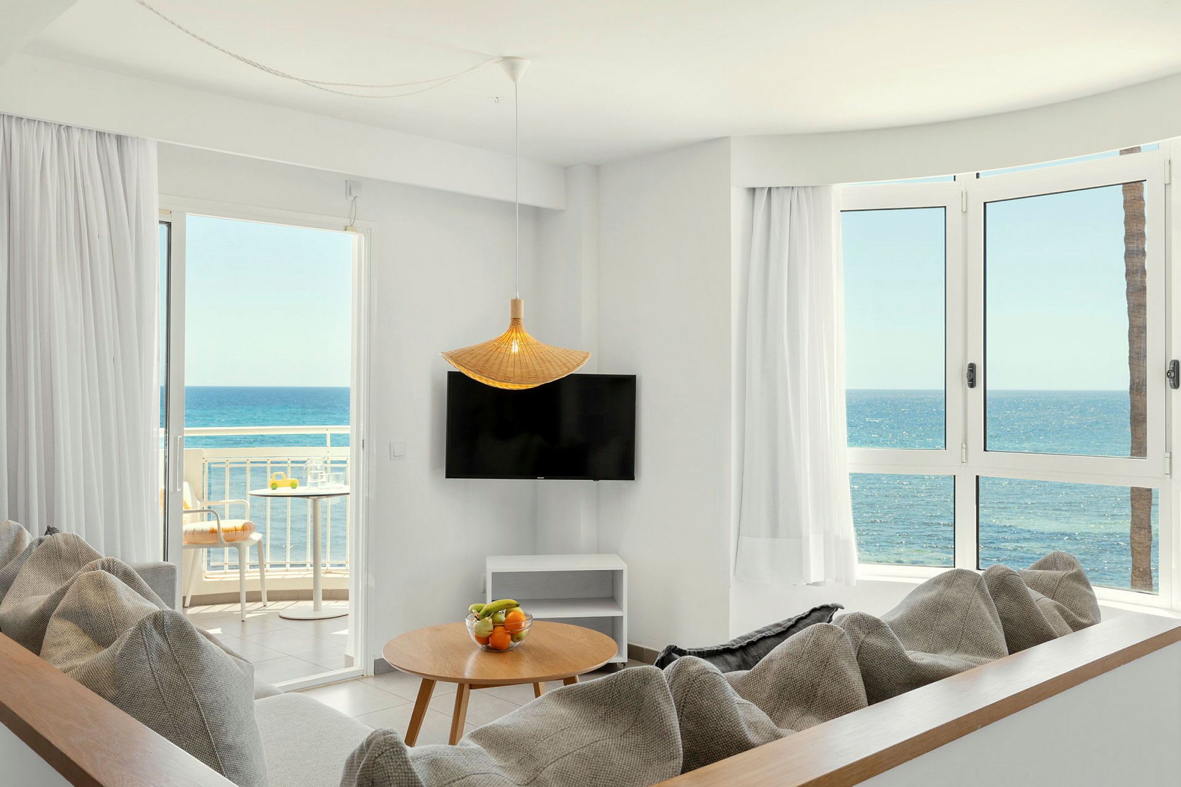 En lys stue med sofaer, et bord med frugtskål, et TV, store vinduer og en balkon med udsigt over havet. (AI-genereret tekst)