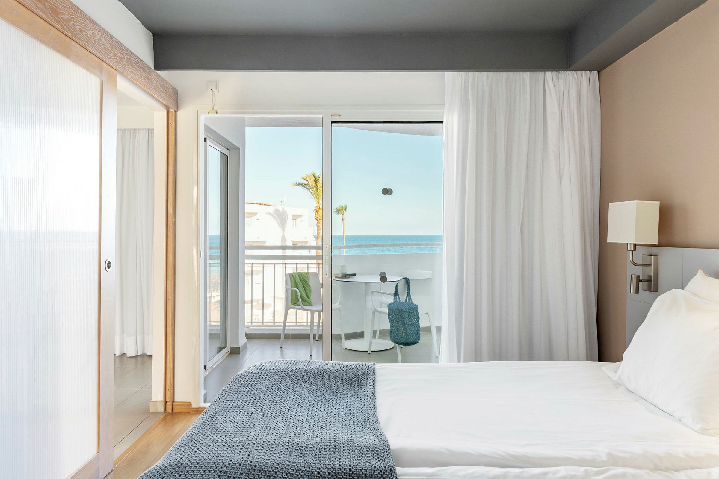 Hotelværelse med seng tæt på en lysåbning mod en balkon med havudsigt. (AI-genereret tekst)