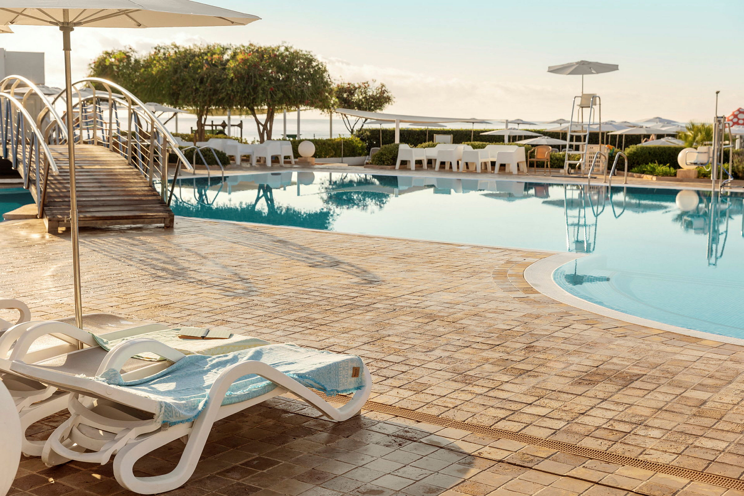 En udendørs swimmingpool med liggestole, parasoller og en bro over vandet. (AI-genereret tekst)