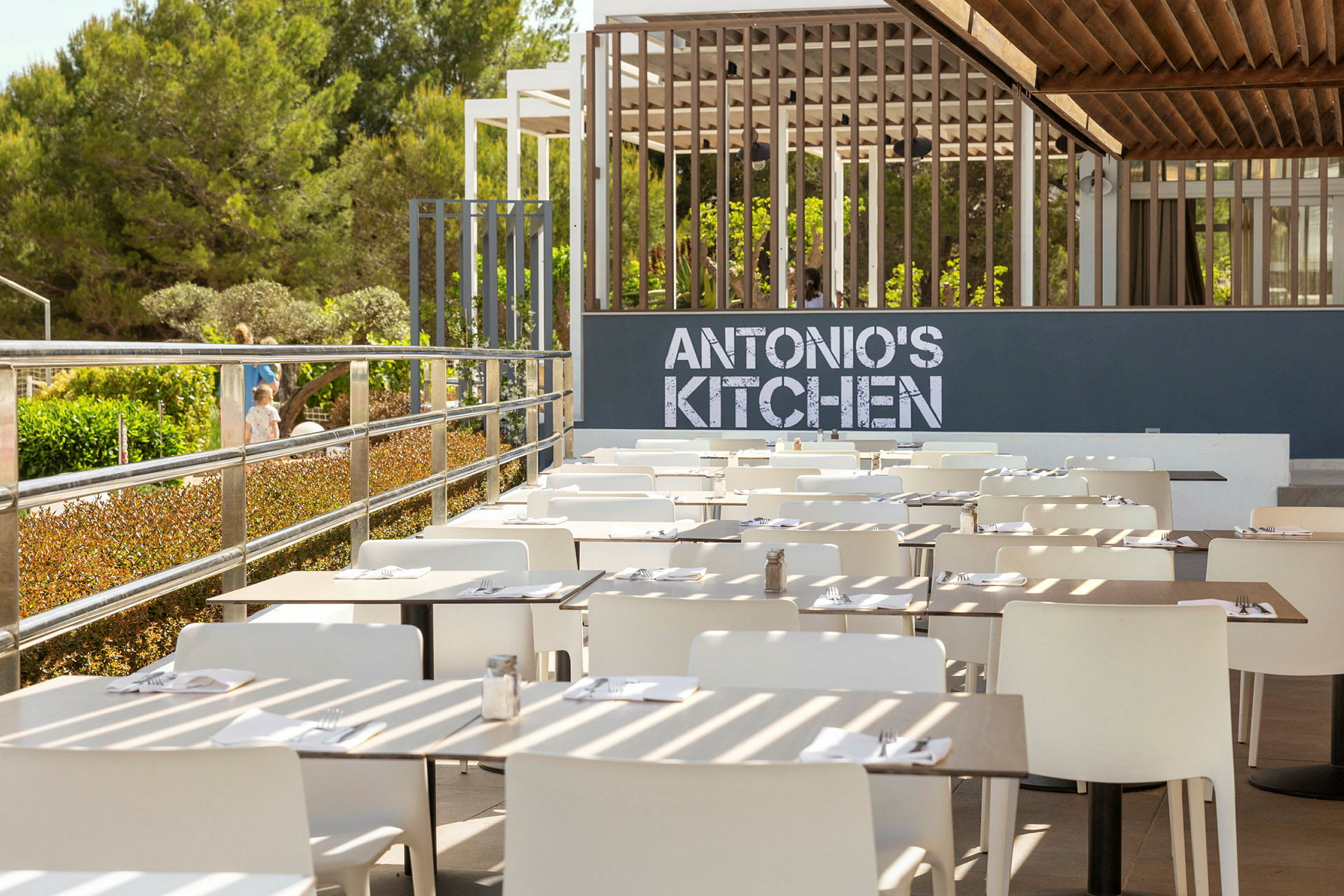 En udendørs terrasse med hvide stole og dækkede borde foran et skilt med teksten 'ANTONIO'S KITCHEN'. (AI-genereret tekst)