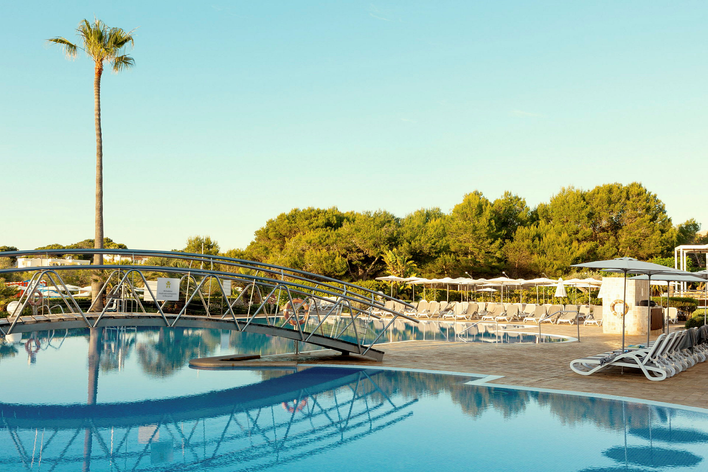 Pool med en bro i forgrunden, liggestole og parasoller rundt omkring samt træer i baggrunden. (AI-genereret tekst)