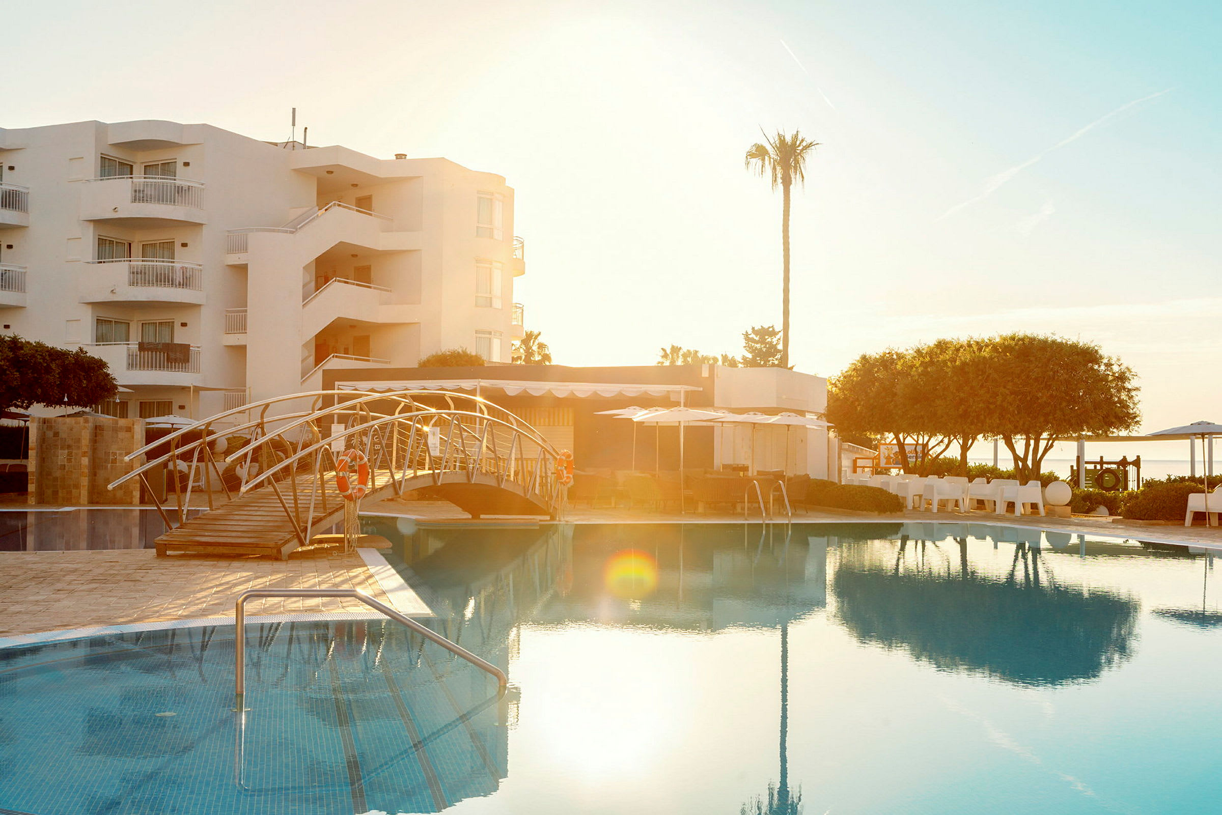 Billede af en udendørs swimmingpool ved et hotel med en bro over poolen, omgivet af grønne områder og bygninger. (AI-genereret tekst)