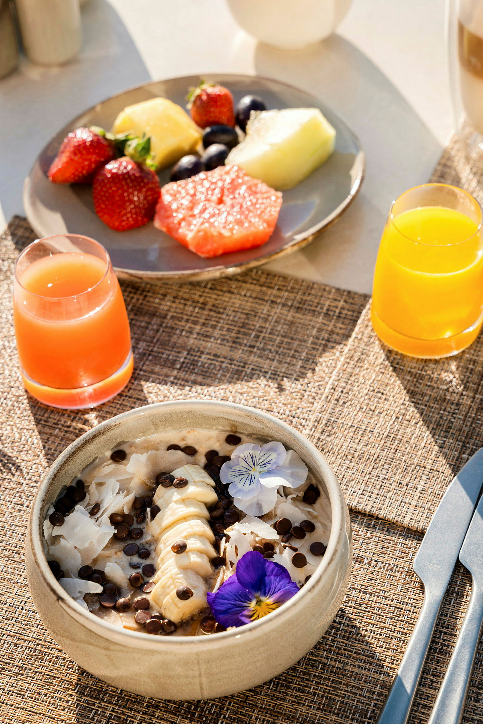 Morgenbord med frisk frugt, en skål med bananskiver og chokoladestykker samt juice. (AI-genereret tekst)