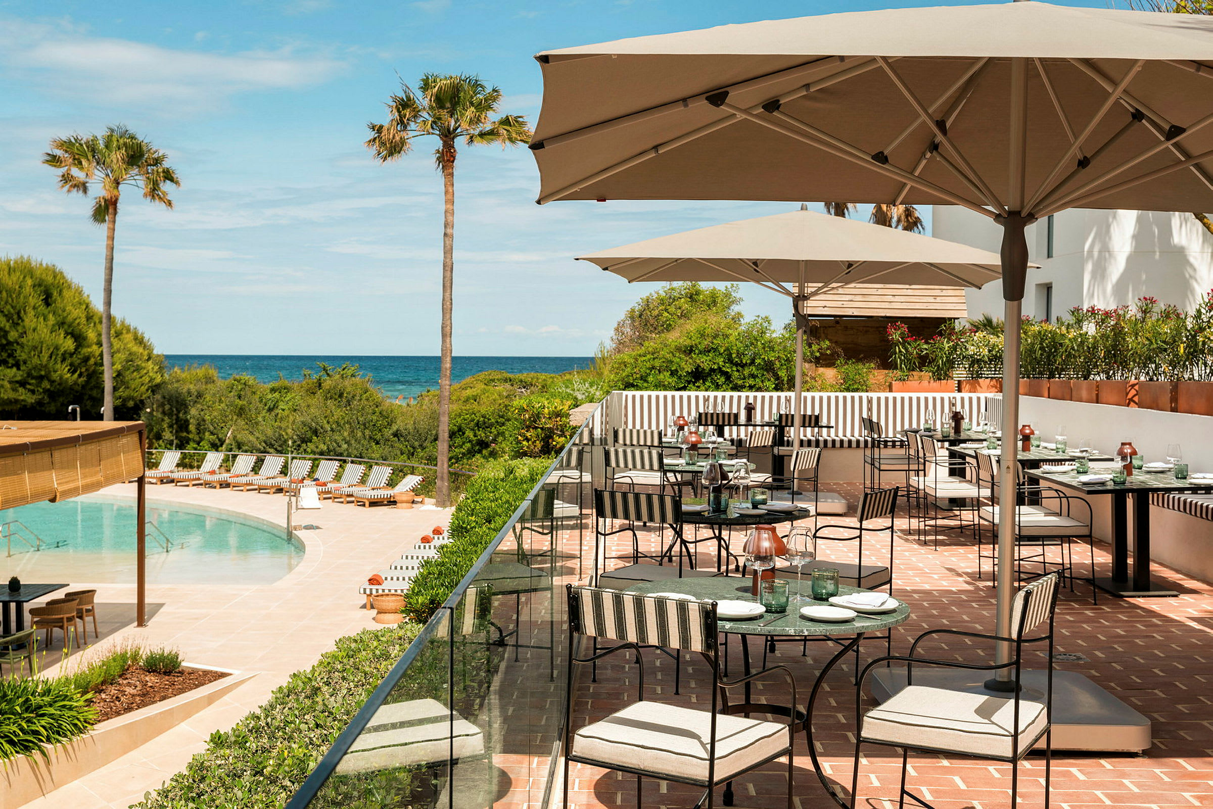 En udendørs terrasse ved en pool med siddegrupper og parasoller. I baggrunden ses palmer og havet. (AI-genereret tekst)