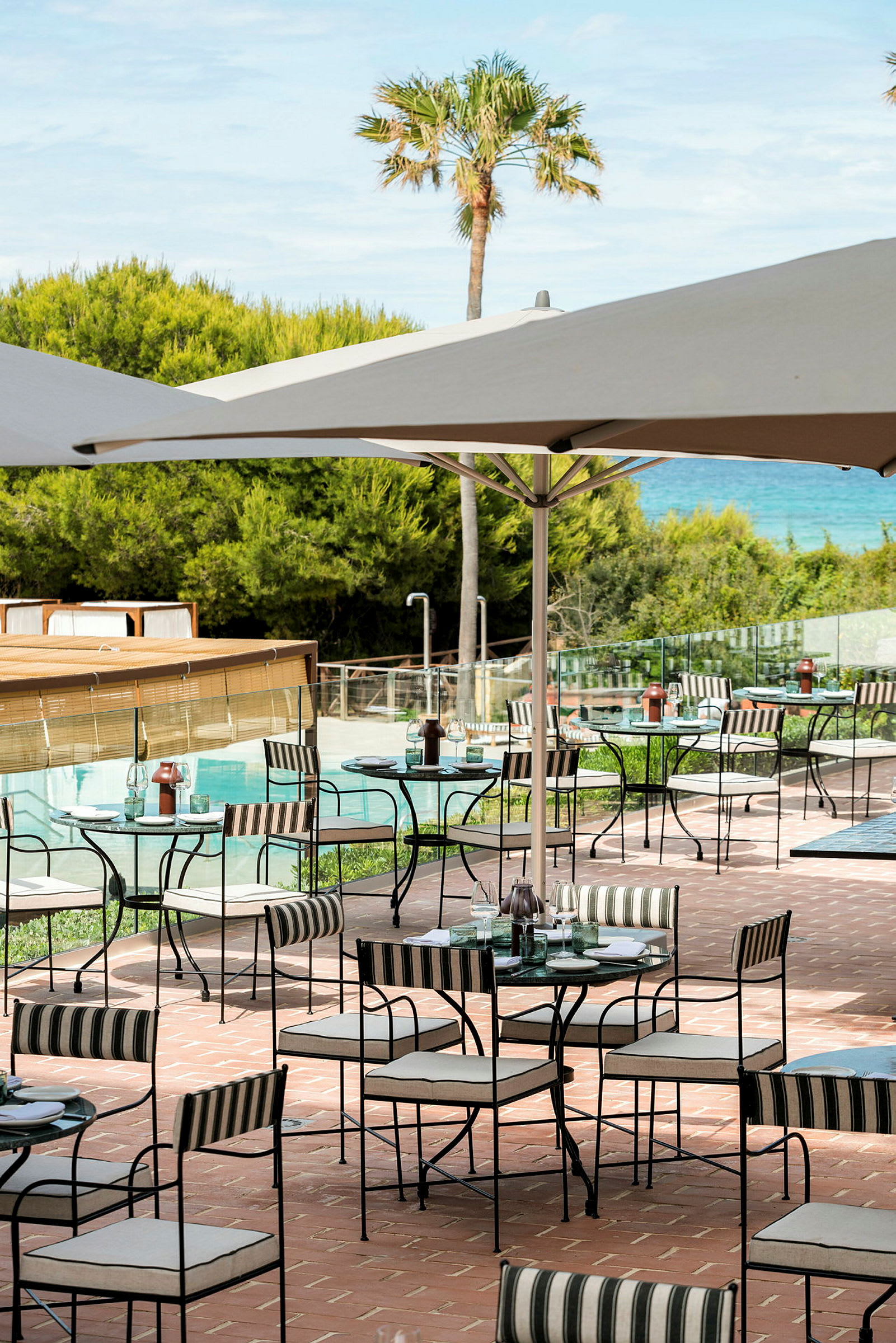 En udendørsservering med borde og stole under parasoller på en terrasse med udsigt over grønne områder og en pool. (AI-genereret tekst)