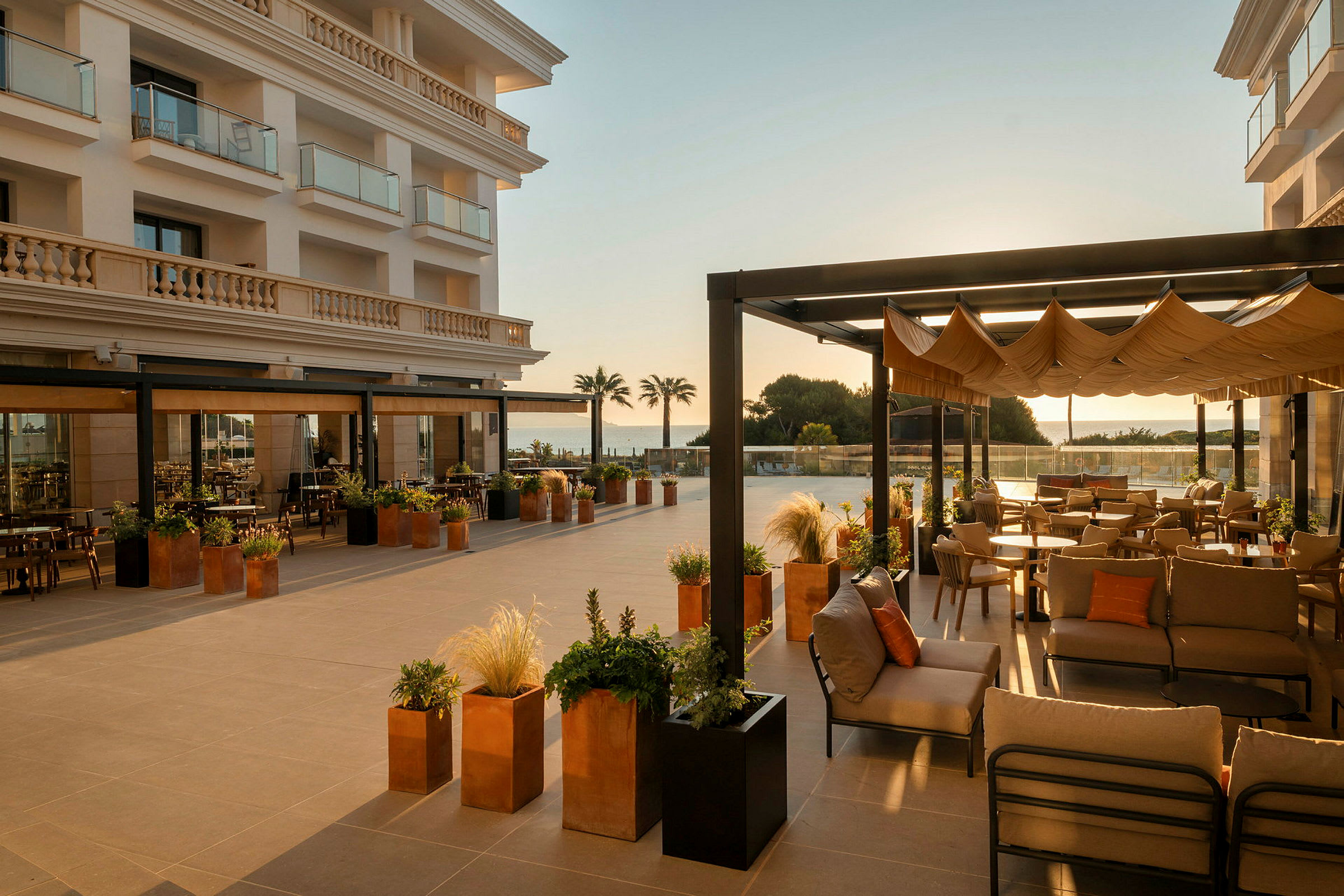 En rummelig udendørsterrasse ved et hotel er indrettet med komfortable møbler og tilbyder udsigt over havet. Detaljer som planter i potter og pergolaer forstærker den indbydende stemning. (AI-genereret tekst)