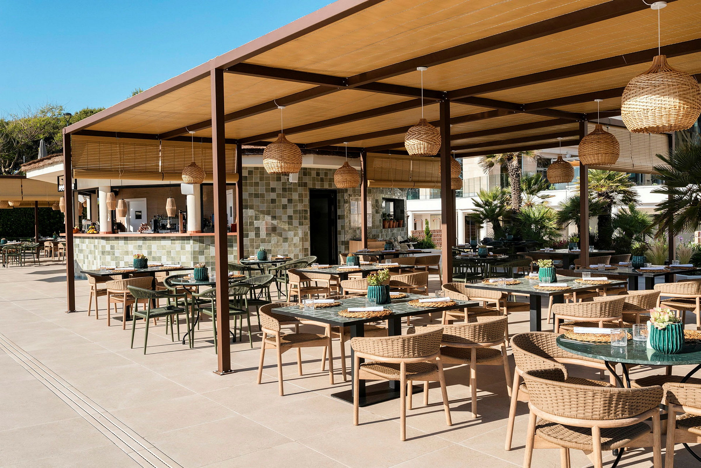 En udendørs restaurant med borde og stole under et tag, med lanterner og en bar i baggrunden. (AI-genereret tekst)