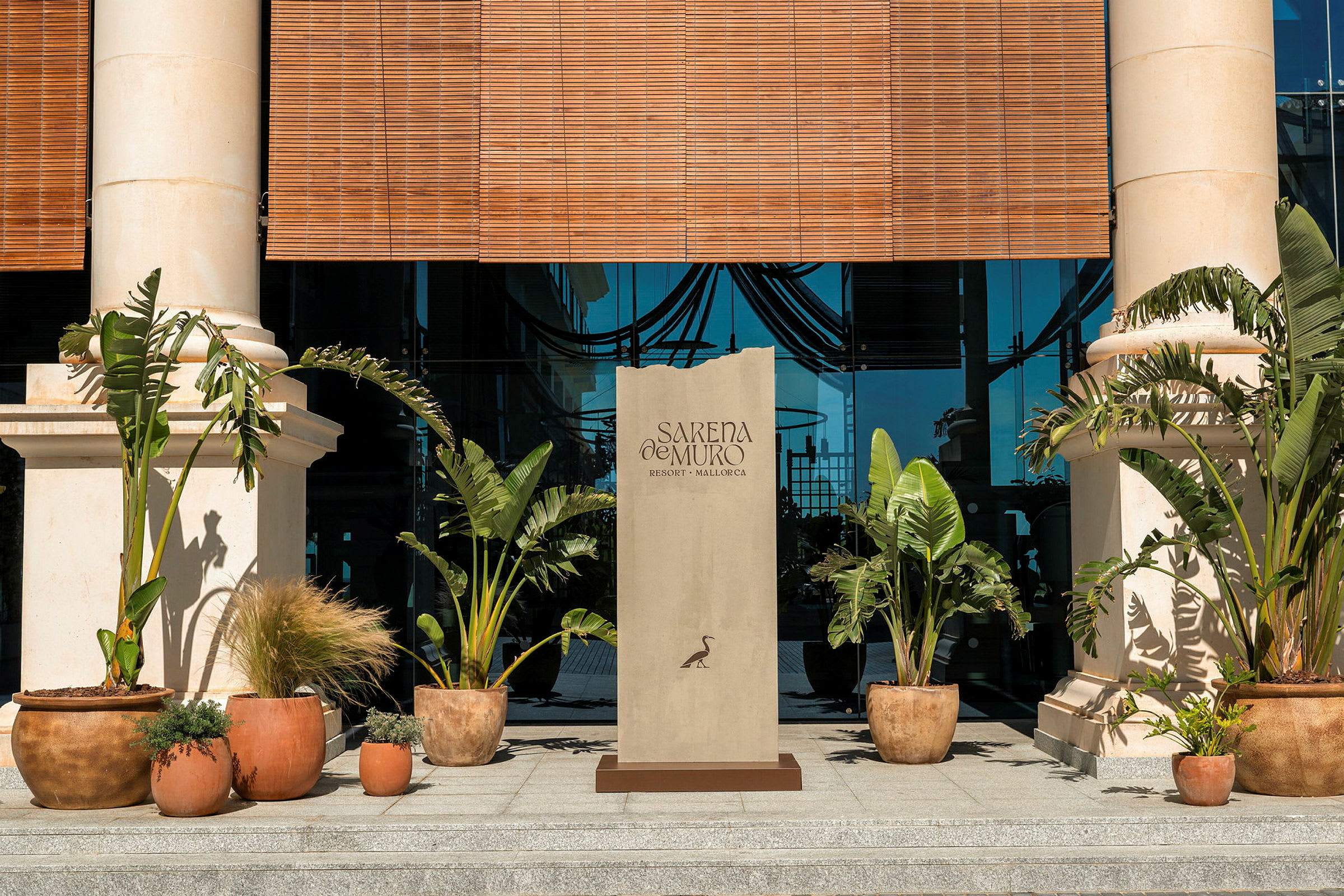 Indgangen til en bygning med to søjler, grønne planter i potter og et skilt med teksten "Sarena de Muro Resort Mallorca". (AI-genereret tekst)