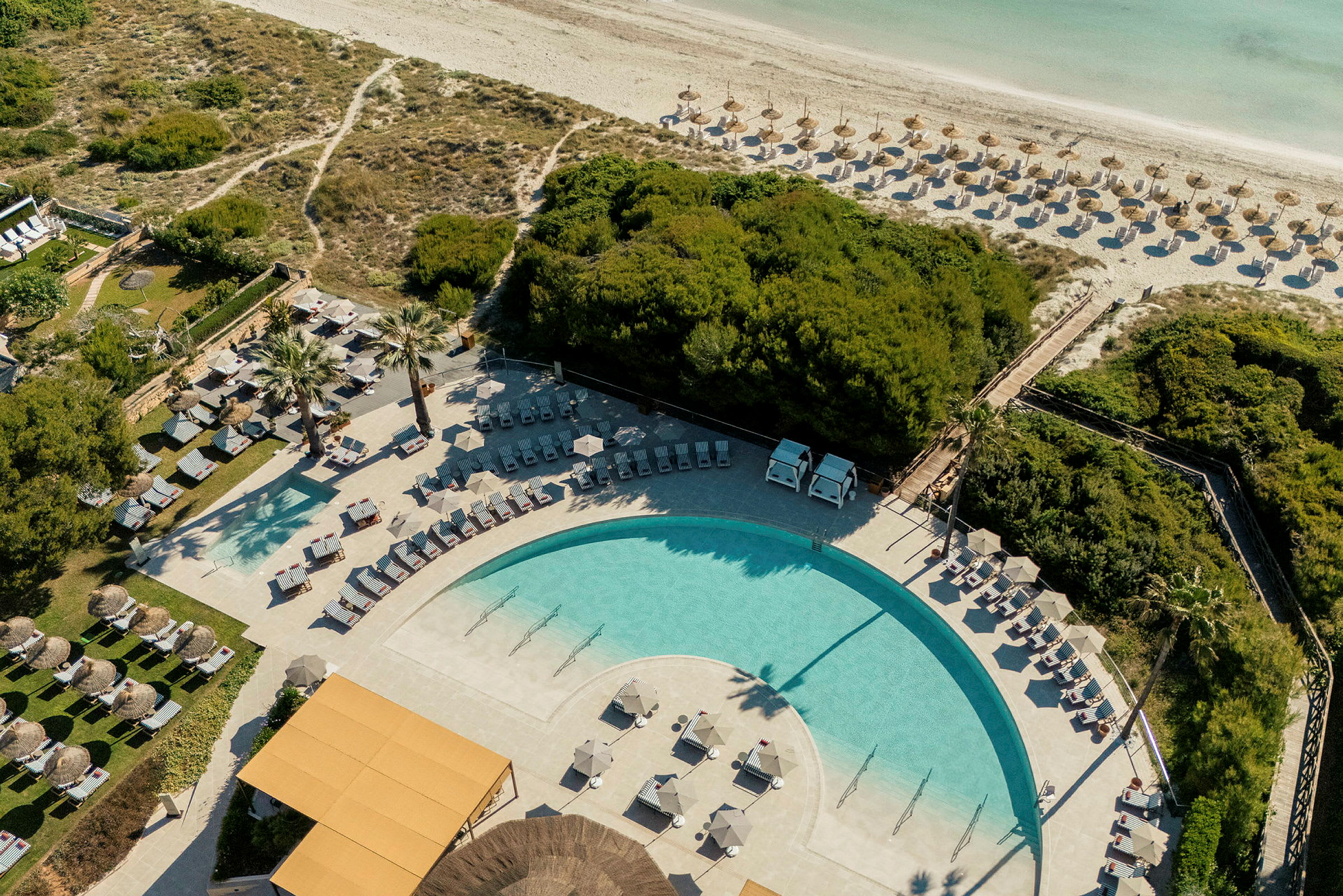 Luftfoto af en pool, liggestole og parasoller omgivet af grønt og en sandstrand i baggrunden. (AI-genereret tekst)