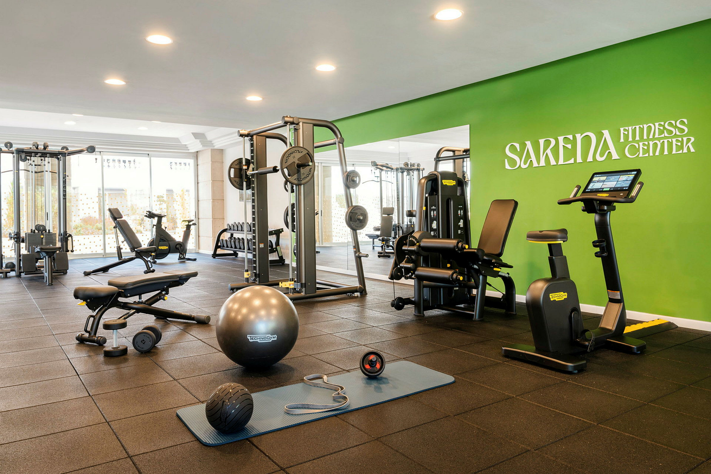 Et fitnesscenter med forskellige træningsmaskiner og redskaber, herunder en kondicykel, håndvægte og en træningsmåtte, samt grøn væg med teksten Sarena Fitness Center. (AI-genereret tekst)
