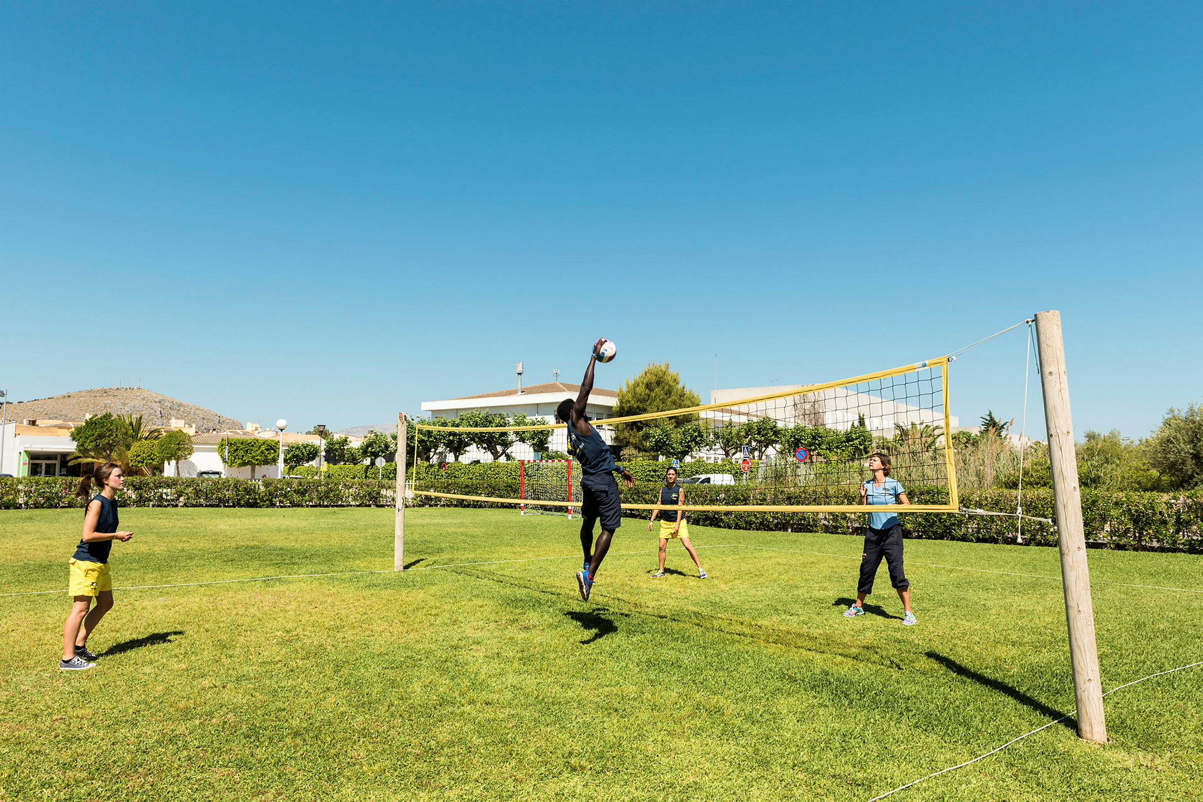 Personer spiller volleyball på en græsplæne under en klar blå himmel. (AI-genereret tekst)