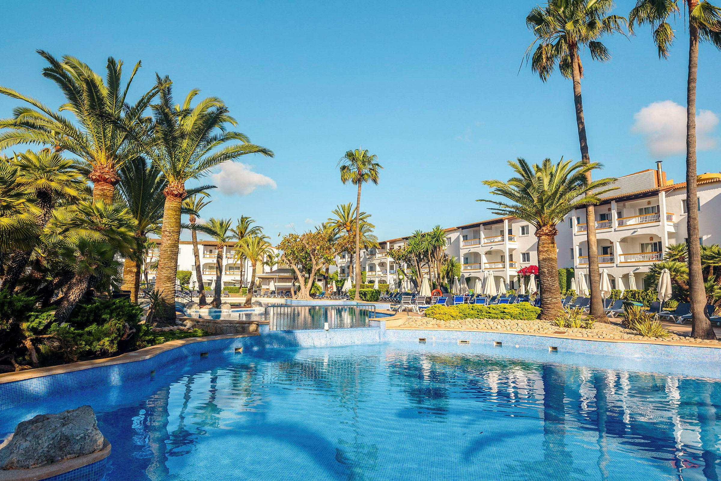 Udendørs pool med palmer og hvidmalet hotelbygning i baggrunden. (AI-genereret tekst)