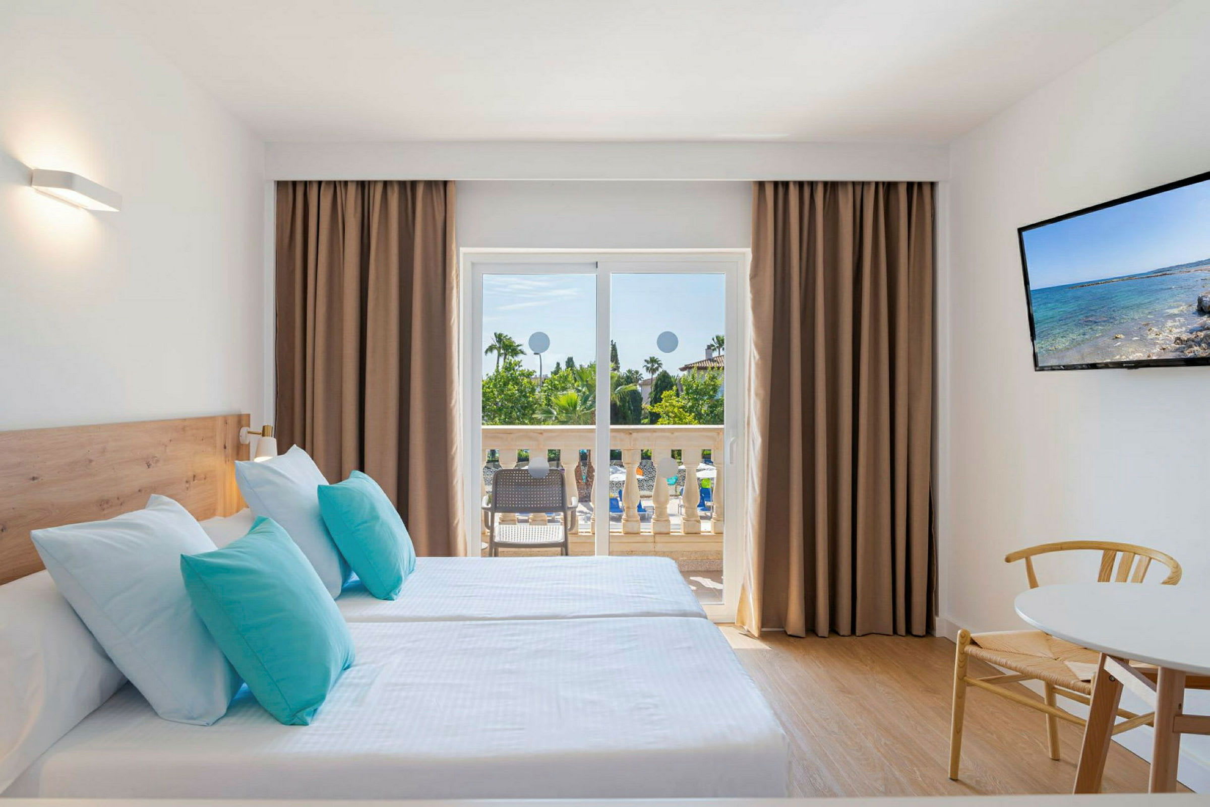 Et hotelværelse med to enkeltsenge med lyseblå og turkise puder, en balkon med udsigt over haven, og et TV på væggen. (AI-genereret tekst)