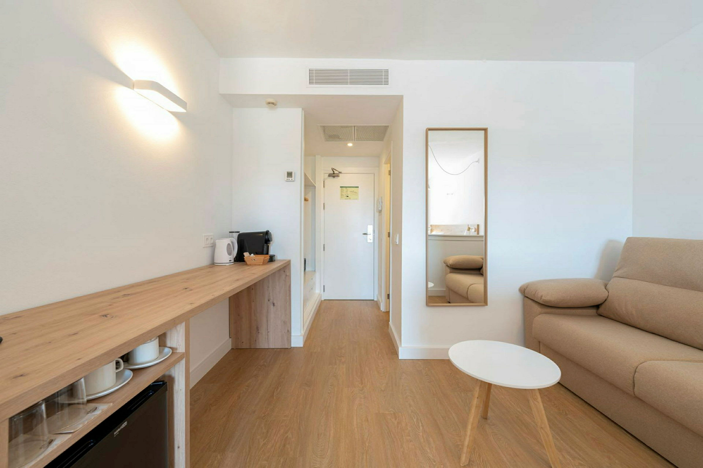 Et moderne hotelværelse med en sofa, spejl, lille bord og en arbejdsplads med elkedel og kaffemaskine. (AI-genereret tekst)