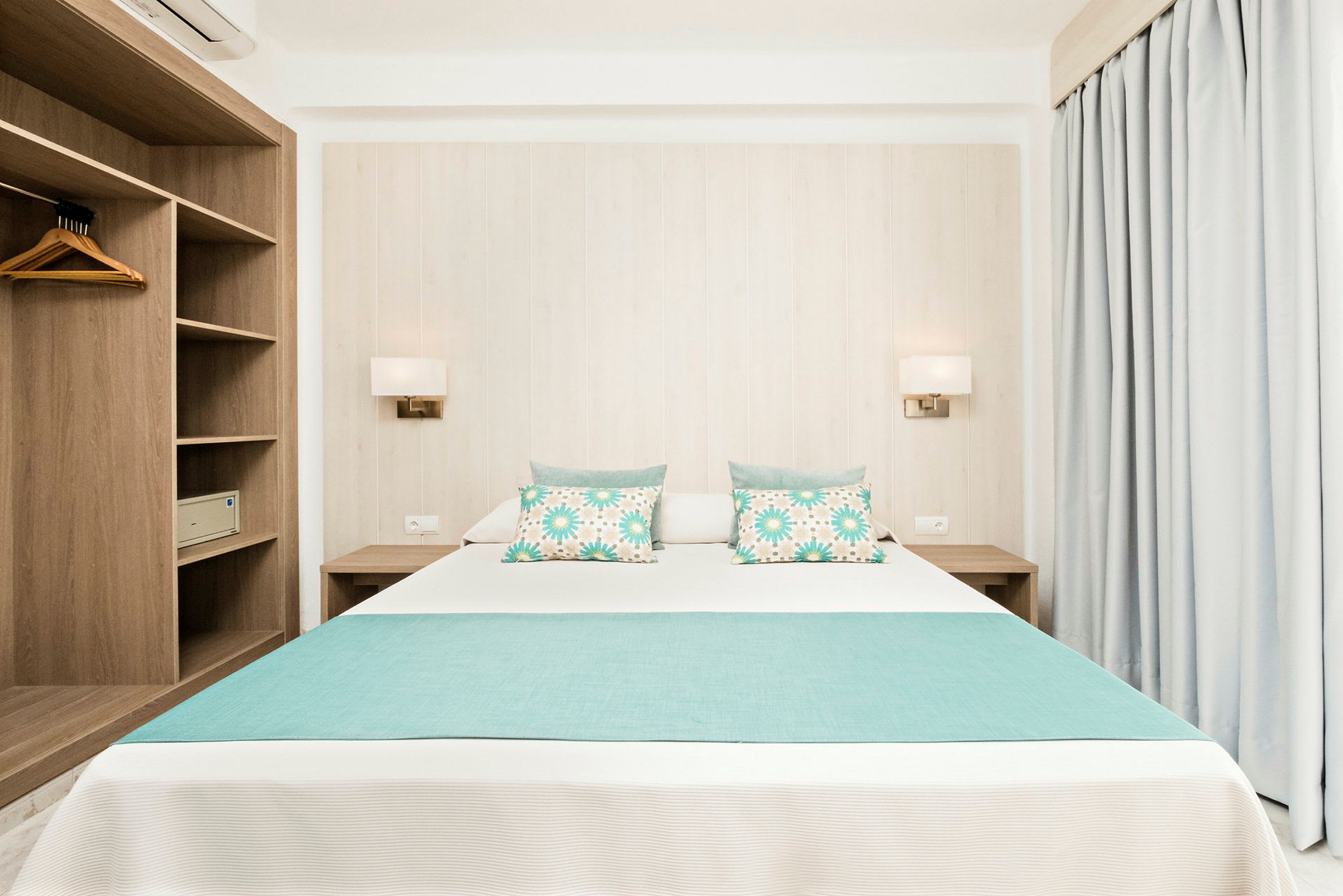 Et lyst og moderne hotelværelse med en dobbeltseng dekoreret med turkise puder og sengetæppe. Til venstre ses et åbent træskab, og til højre er gardiner. (AI-genereret tekst)