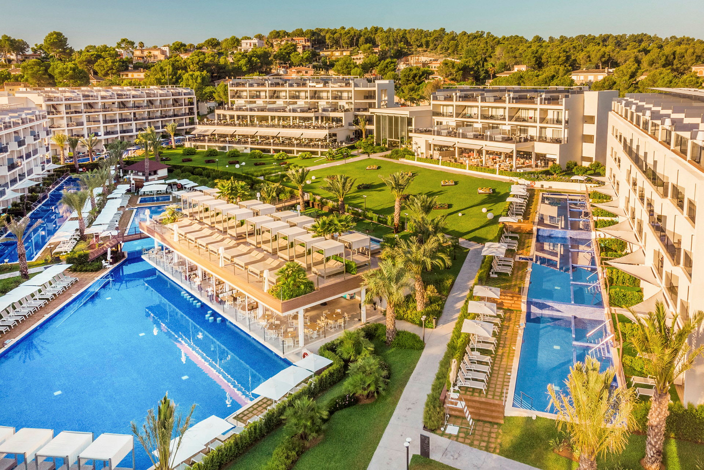 Flere hotelbygninger med store pools og grønne områder omgivet af træer. (AI-genereret tekst)