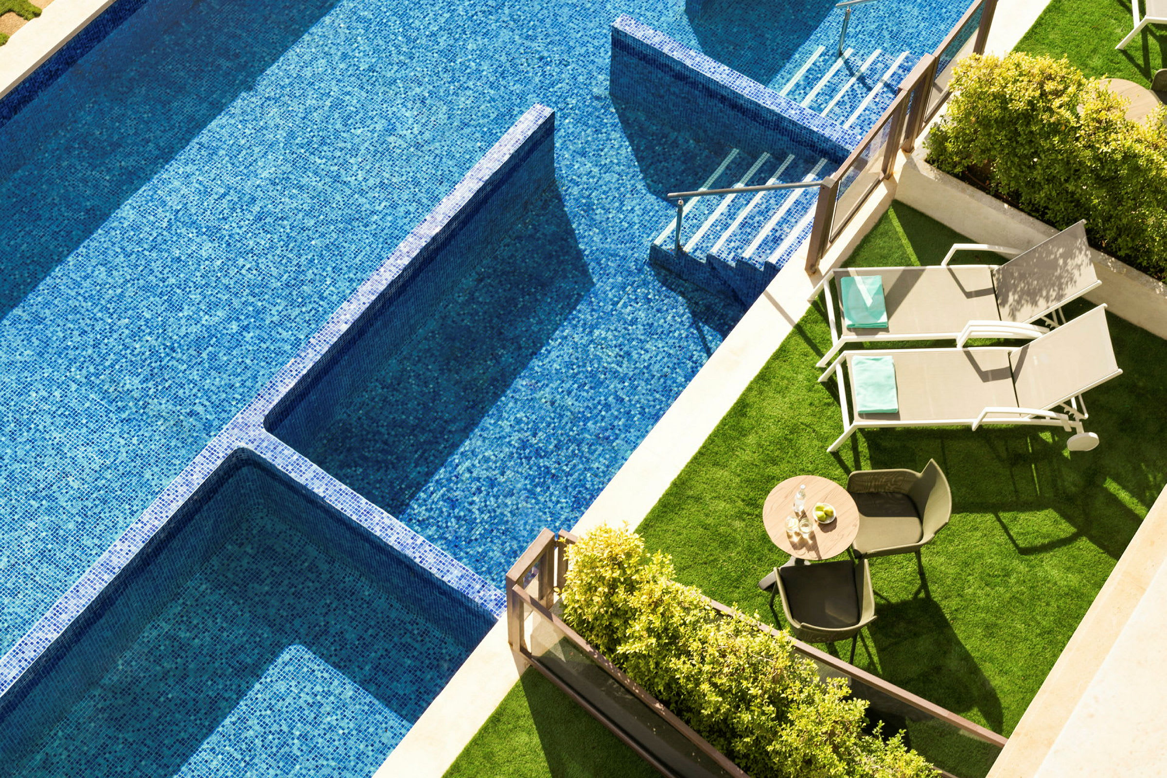 Billede af en swimmingpool med blå mosaik, to liggestole og et bord med stole på et grønt område. (AI-genereret tekst)