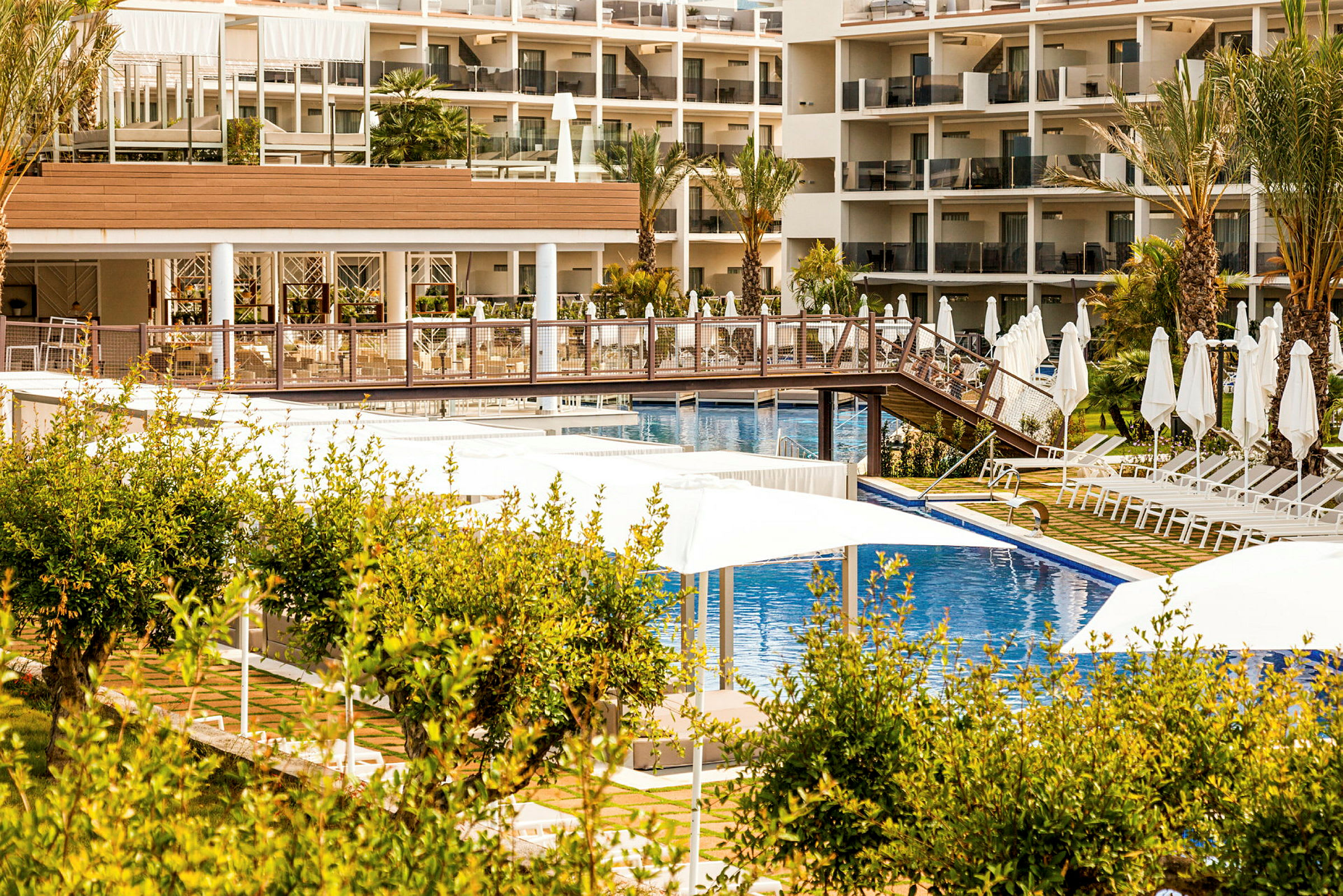 Hotelområde med pool, liggestole og parasoller udendørs, omgivet af grønne områder. (AI-genereret tekst)