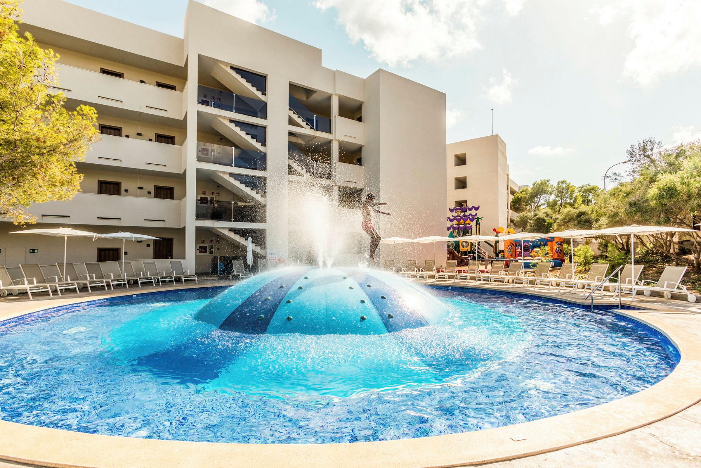 Udendørs pool med springvand tæt på hotelbygning. (AI-genereret tekst)