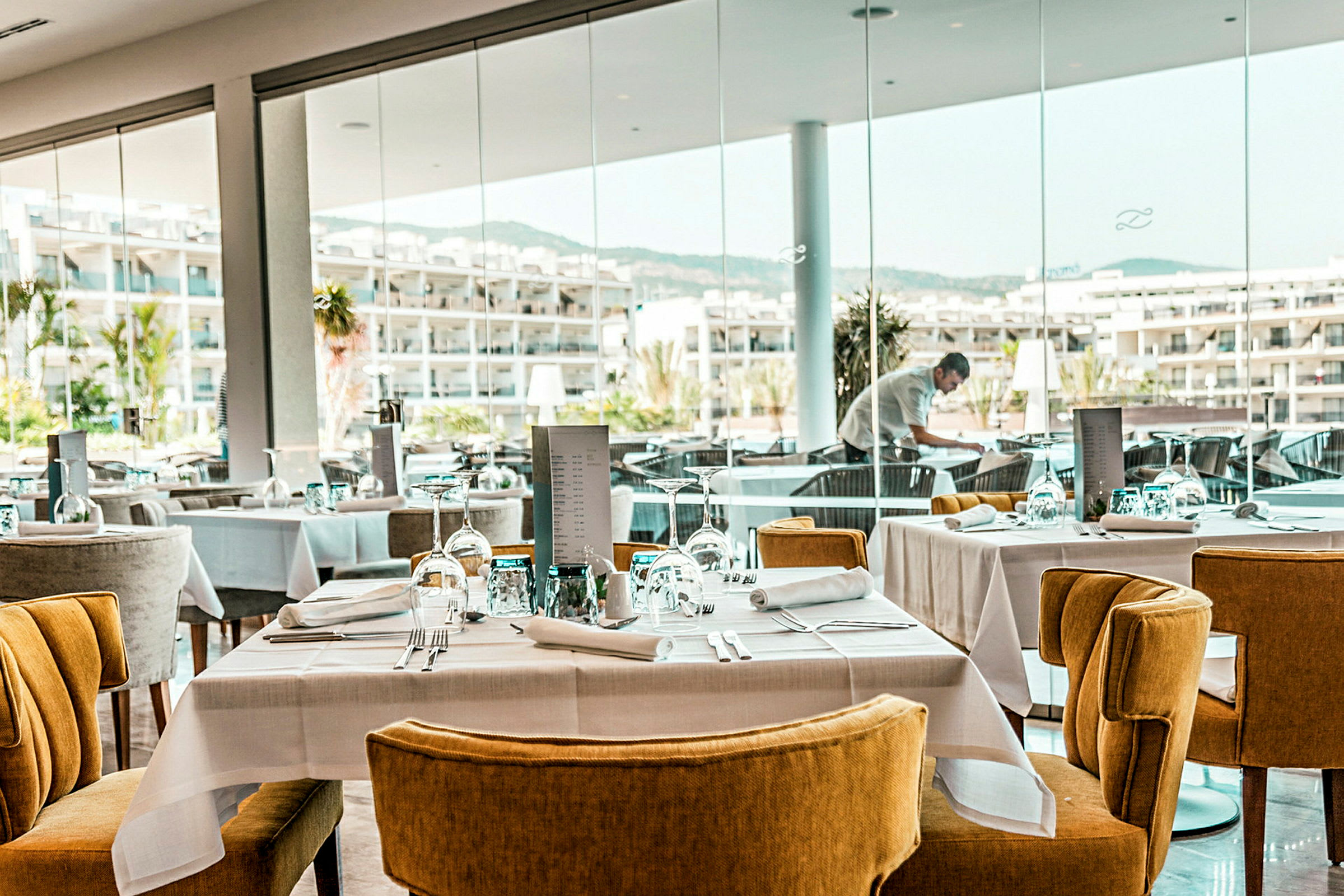 En moderne restaurant med dækkede borde og stole samt vinduer med udsigt over et hotelområde. (AI-genereret tekst)