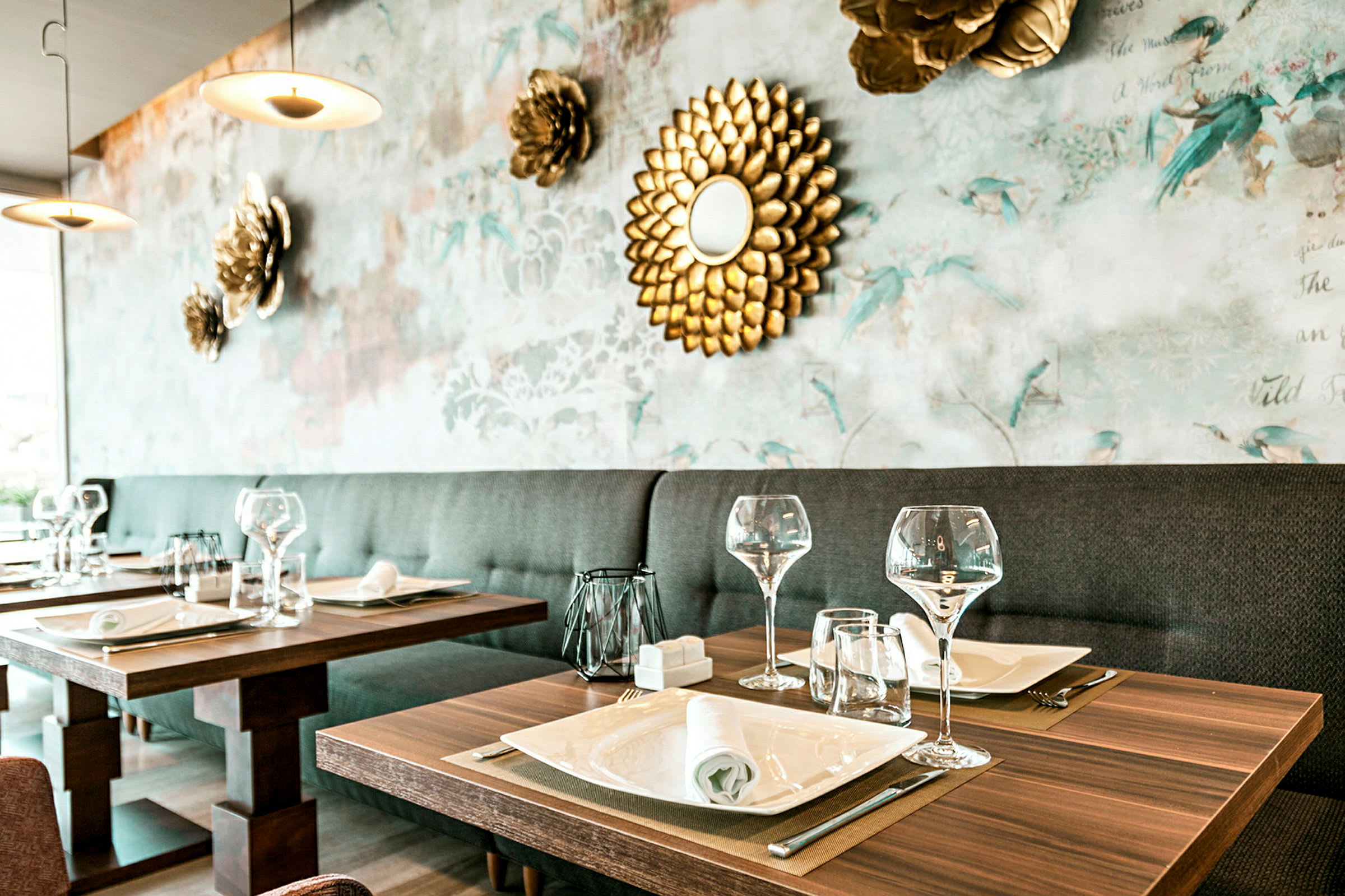 Et elegant restaurantbord dækket med glas, tallerkener og servietter, med en dekorativ baggrunds væg prydet med blomster og et gyldent spejl. (AI-genereret tekst)