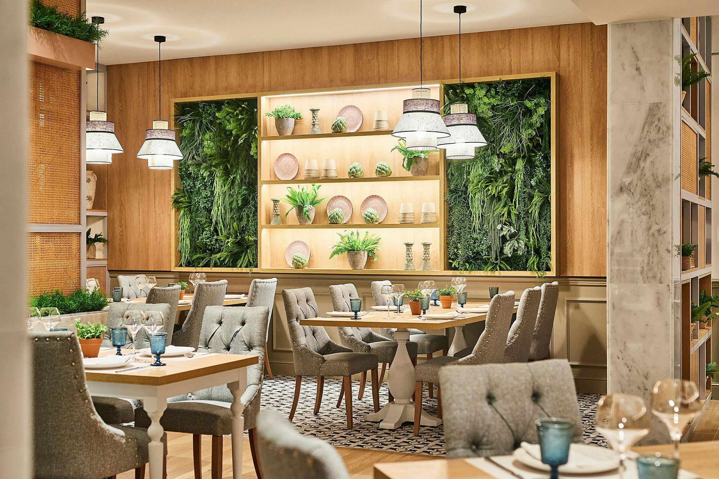 En restaurant med lys og moderne indretning. Bordene er omgivet af grå polstrede stole, og på væggen er der grønne plantepaneler og hylder med dekorationer. (AI-genereret tekst)