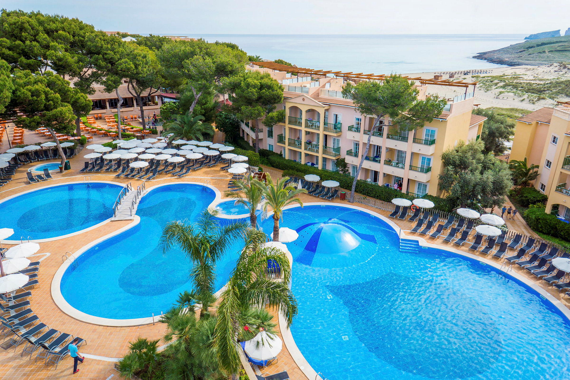 Hotelområde med flere pools, liggestole og parasoller, omgivet af bygninger og træer. (AI-genereret tekst)