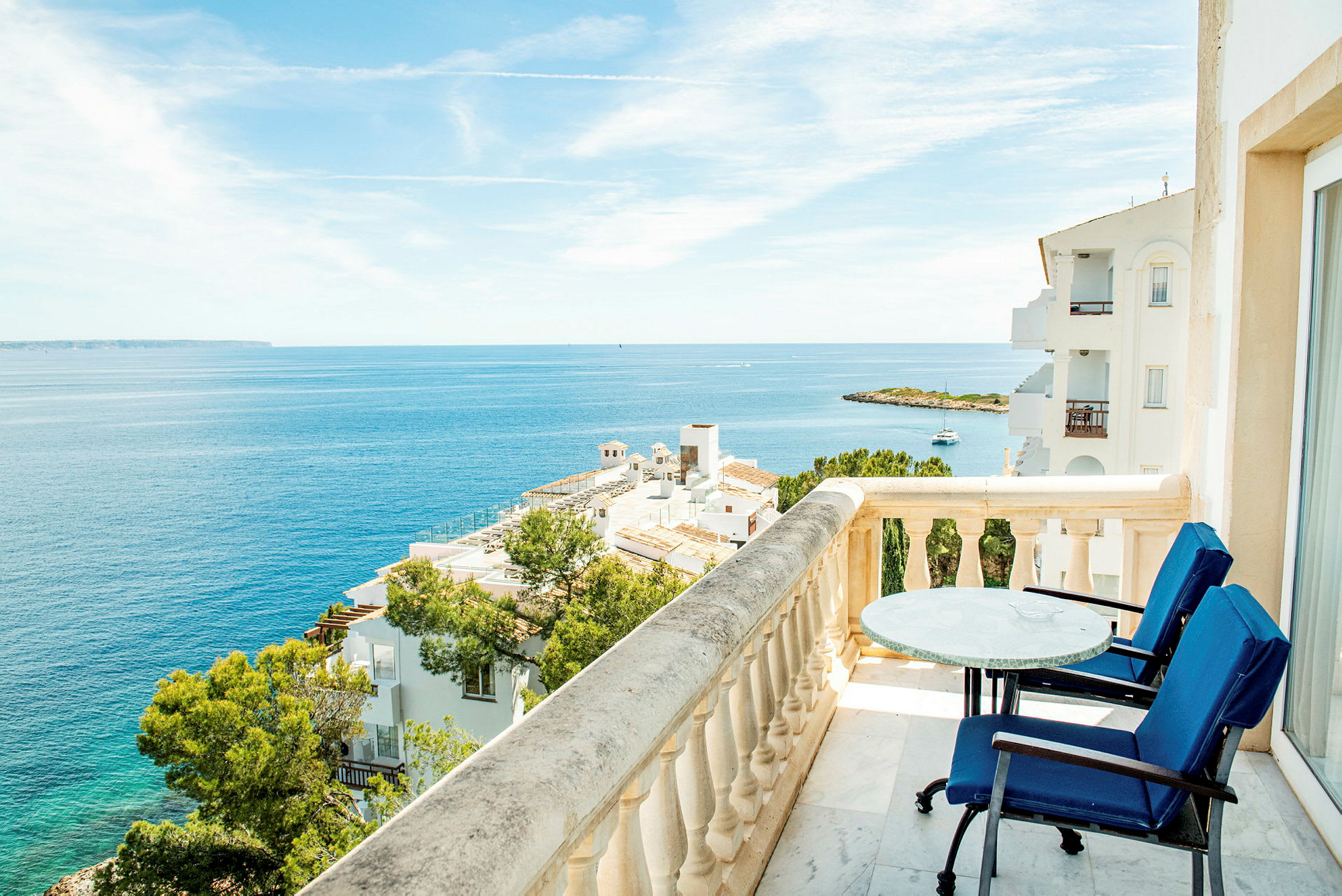 Udsigt fra en balkon med bord og stole over havet og omkringliggende bygninger. (AI-genereret tekst)