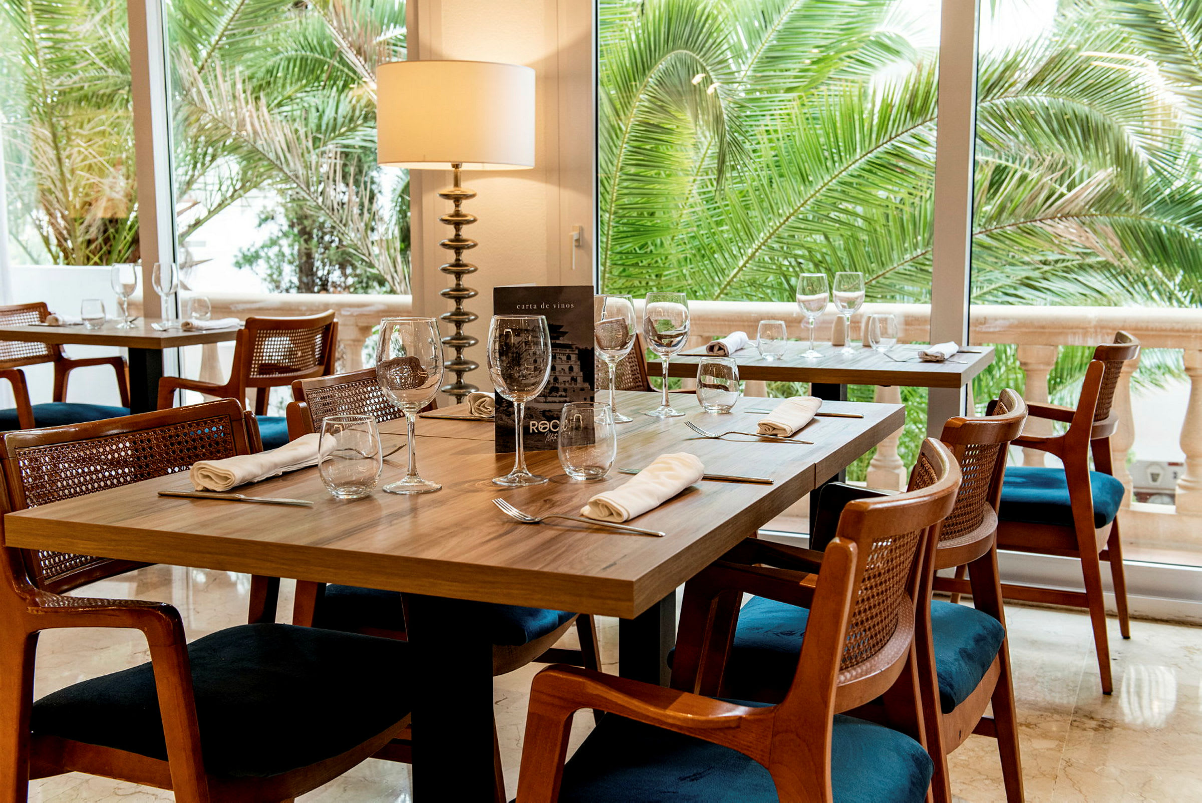 Et restaurantbord med opdækning og glas, omgivet af træstole og grønne palmer synlige gennem vinduet. (AI-genereret tekst)