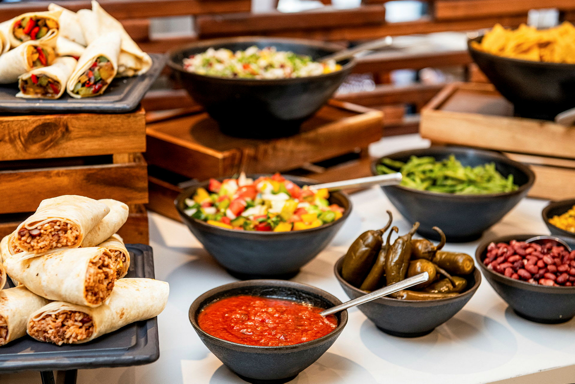 Billede af en buffet med burritos, salat, jalapeños, sauce og andre tilbehør. (AI-genereret tekst)