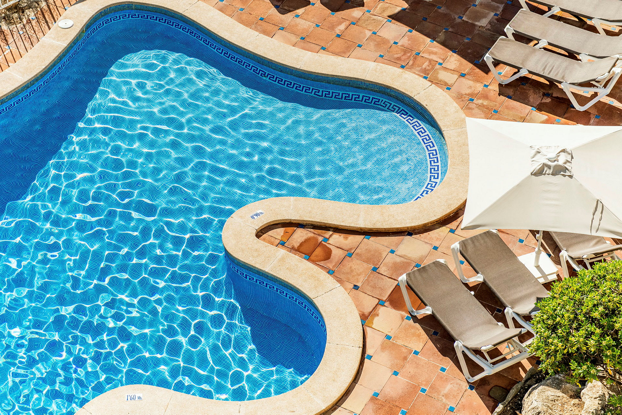 En formet swimmingpool med blåt vand og omkransende solsenge på stenbelagt overflade. (AI-genereret tekst)