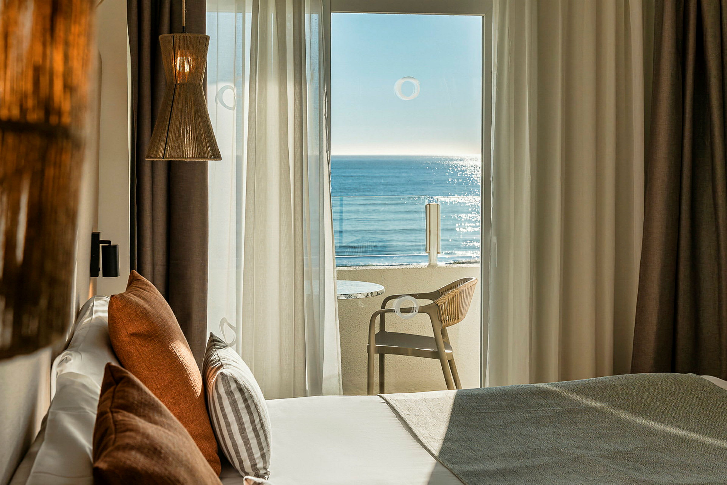 Et hotelværelse med en stor altandør, der fører til en altan med udsigt over havet. Sengen er udstyret med dekorative puder, og en lampe hænger fra loftet. (AI-genereret tekst)