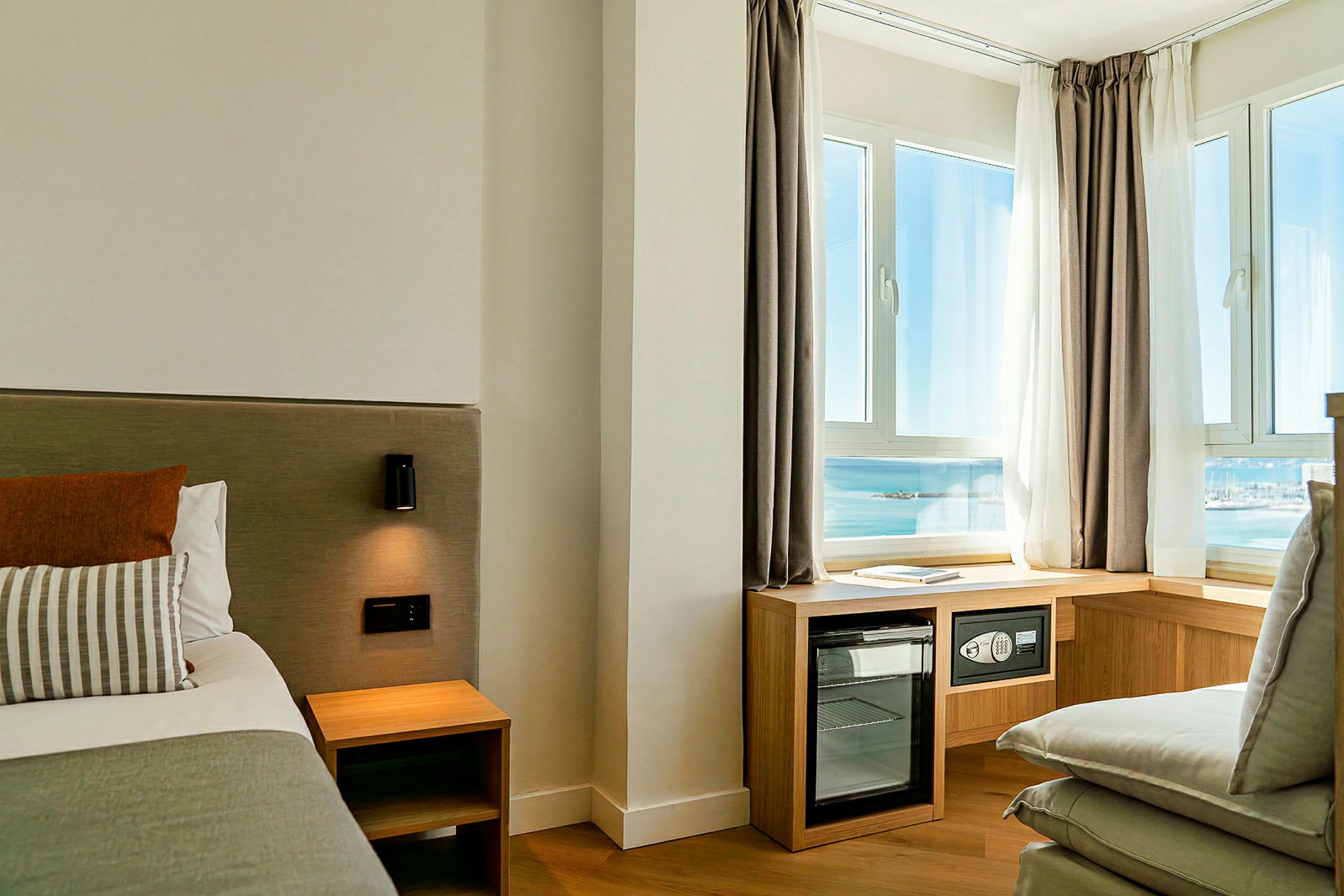 Hotelværelse med en seng, natbord, væglampe og store vinduer med udsigt over havet samt møbler såsom en minibar. (AI-genereret tekst)