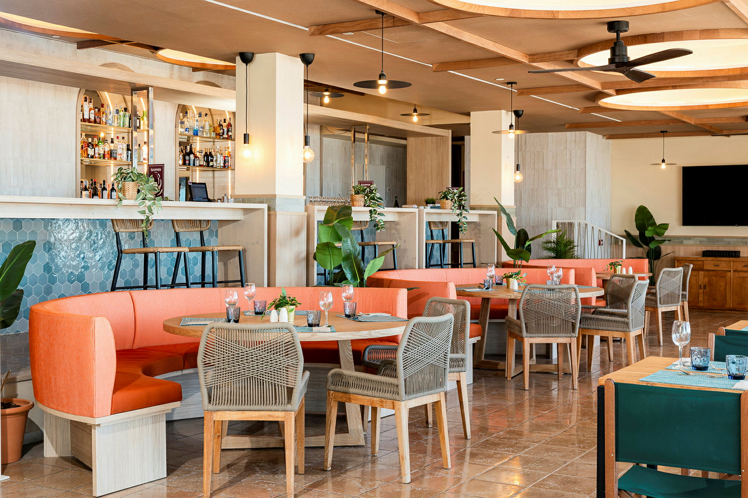 En moderne restaurant med indbydende indretning, orange sofaer og borde med grå stole, samt en bardisk og grønne planter. (AI-genereret tekst)