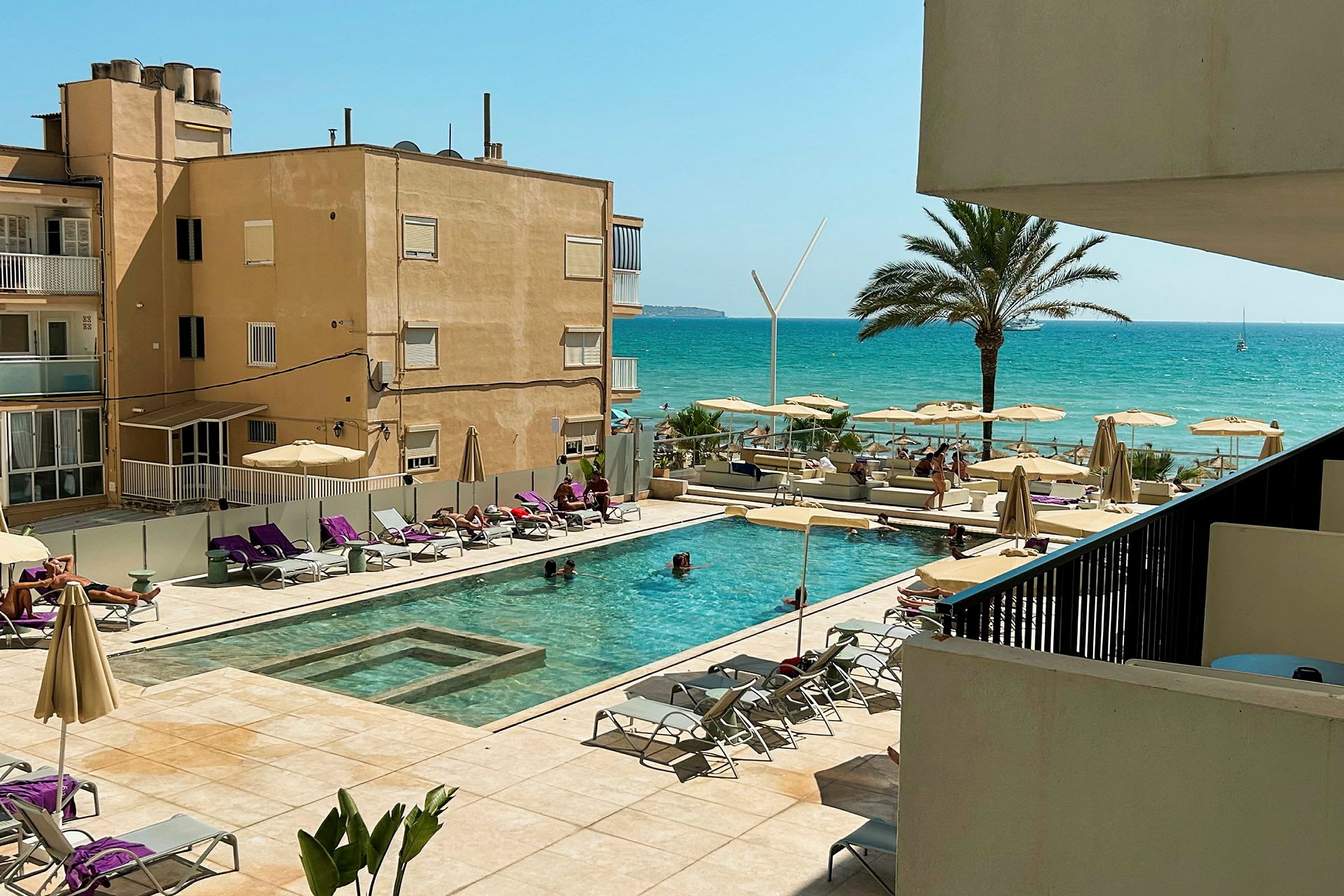 Udsigt over en hotelpool med liggestole og parasoller, med en strand og havet i baggrunden. (AI-genereret tekst)