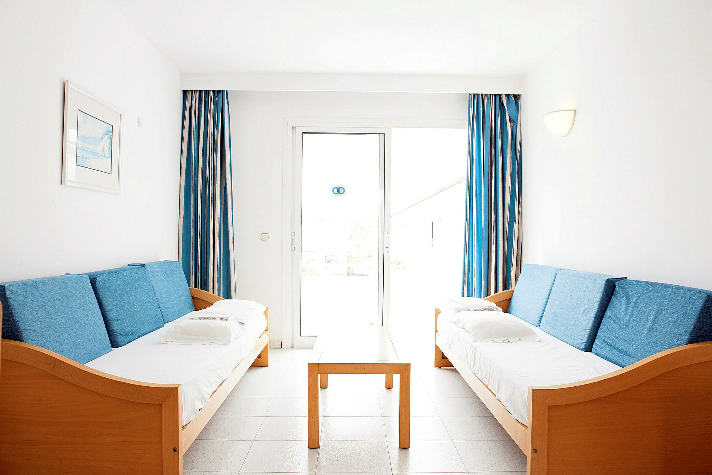 To sofaer med blå puder står over for hinanden, med et træbord imellem og blå-hvide gardiner ved vinduet i baggrunden. (AI-genereret tekst)