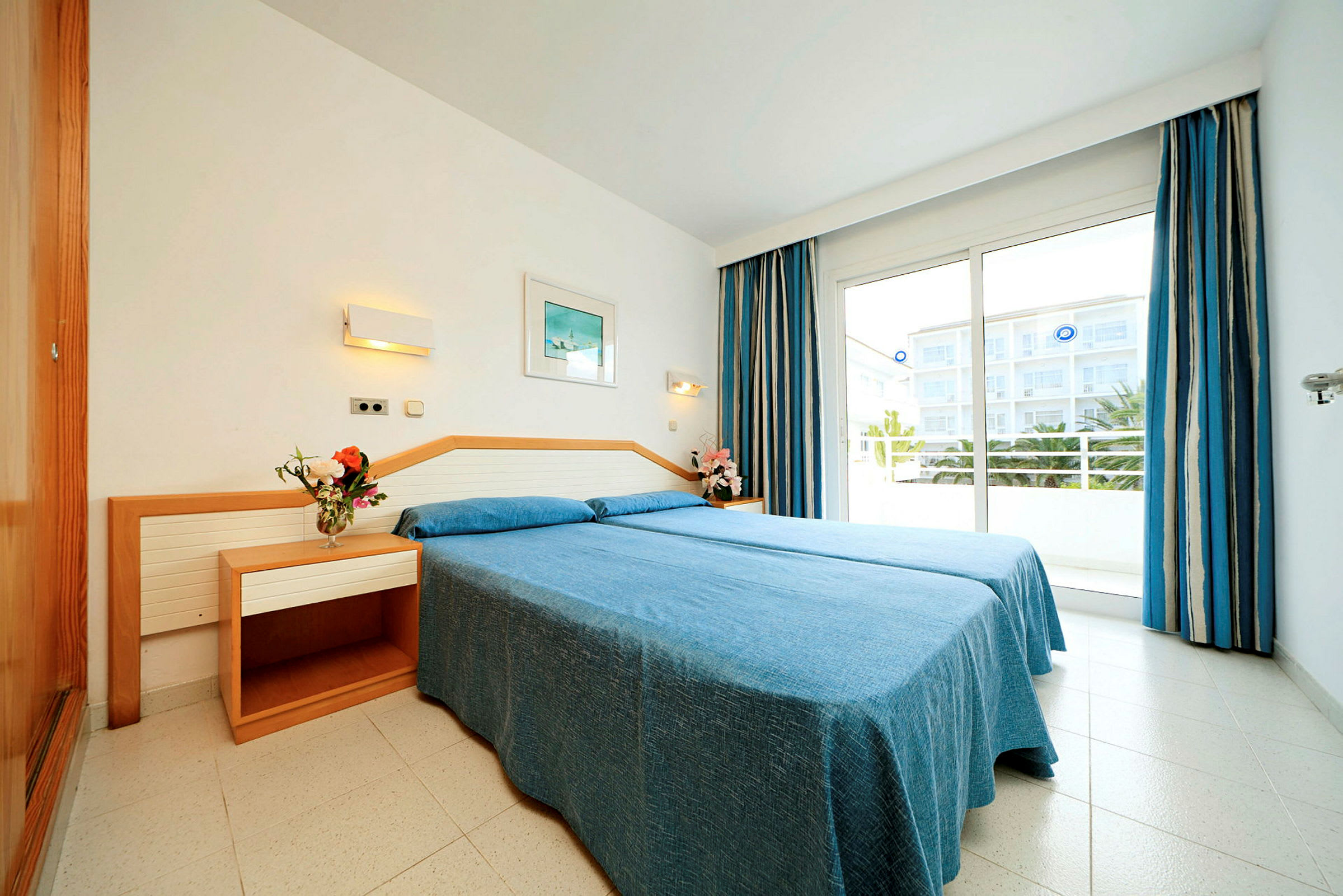 Et lyst hotelværelse med en dobbeltseng med blå sengelinned, to natborde med dekorationer og en balkon med udsigt. (AI-genereret tekst)