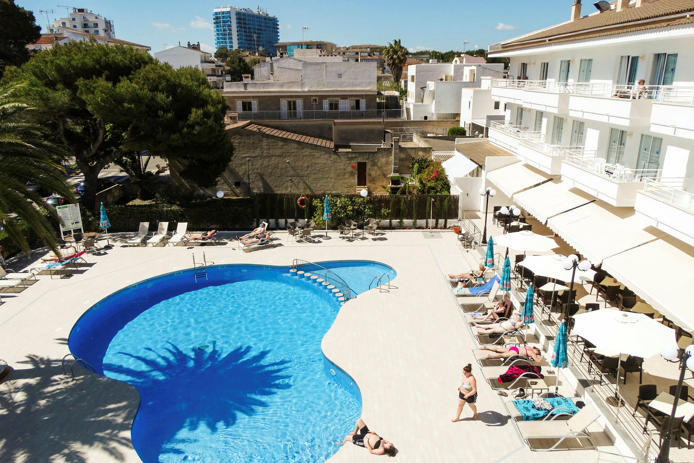 Udendørs pool med liggestole og parasoller ved siden af, flankeret af et hotel og omkringliggende bygninger. (AI-genereret tekst)