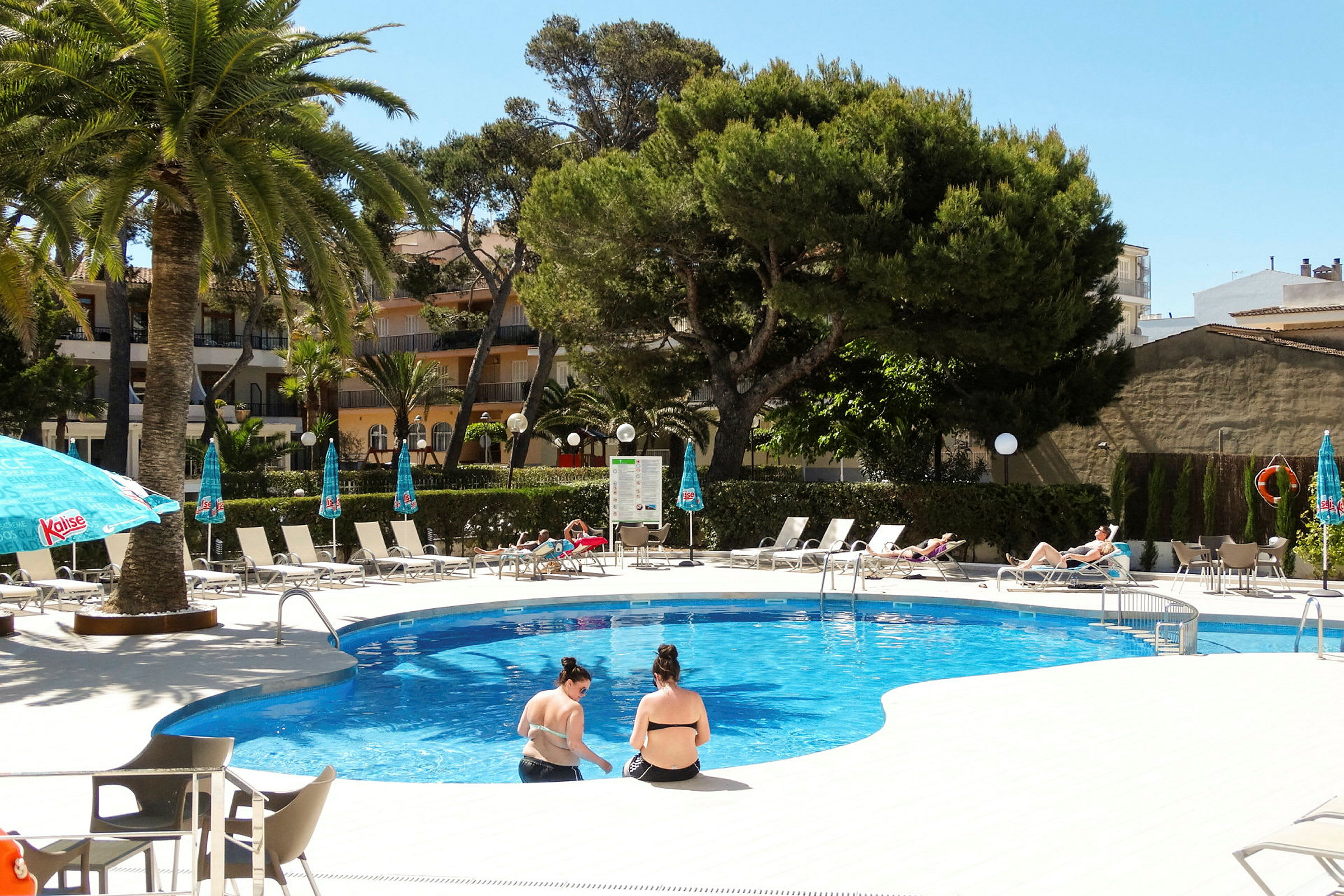 Billede af en udendørs swimmingpool omgivet af liggestole og parasoller i en frodig omgivelser. (AI-genereret tekst)