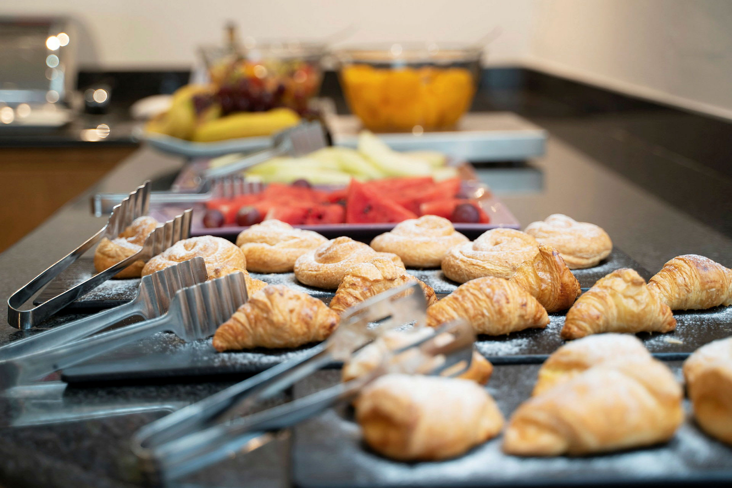 Et buffetbord med croissanter og andre bagværk opstillet ved siden af tænger til servering. Frisk frugt ses i baggrunden. (AI-genereret tekst)