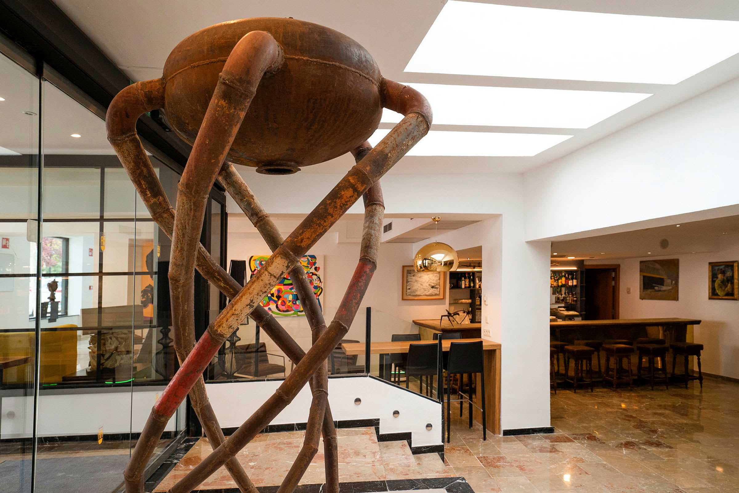 Interiør med en moderne skulptur lavet af rustfarvede metalrør. I baggrunden ses en bar og forskellige kunstværker. (AI-genereret tekst)