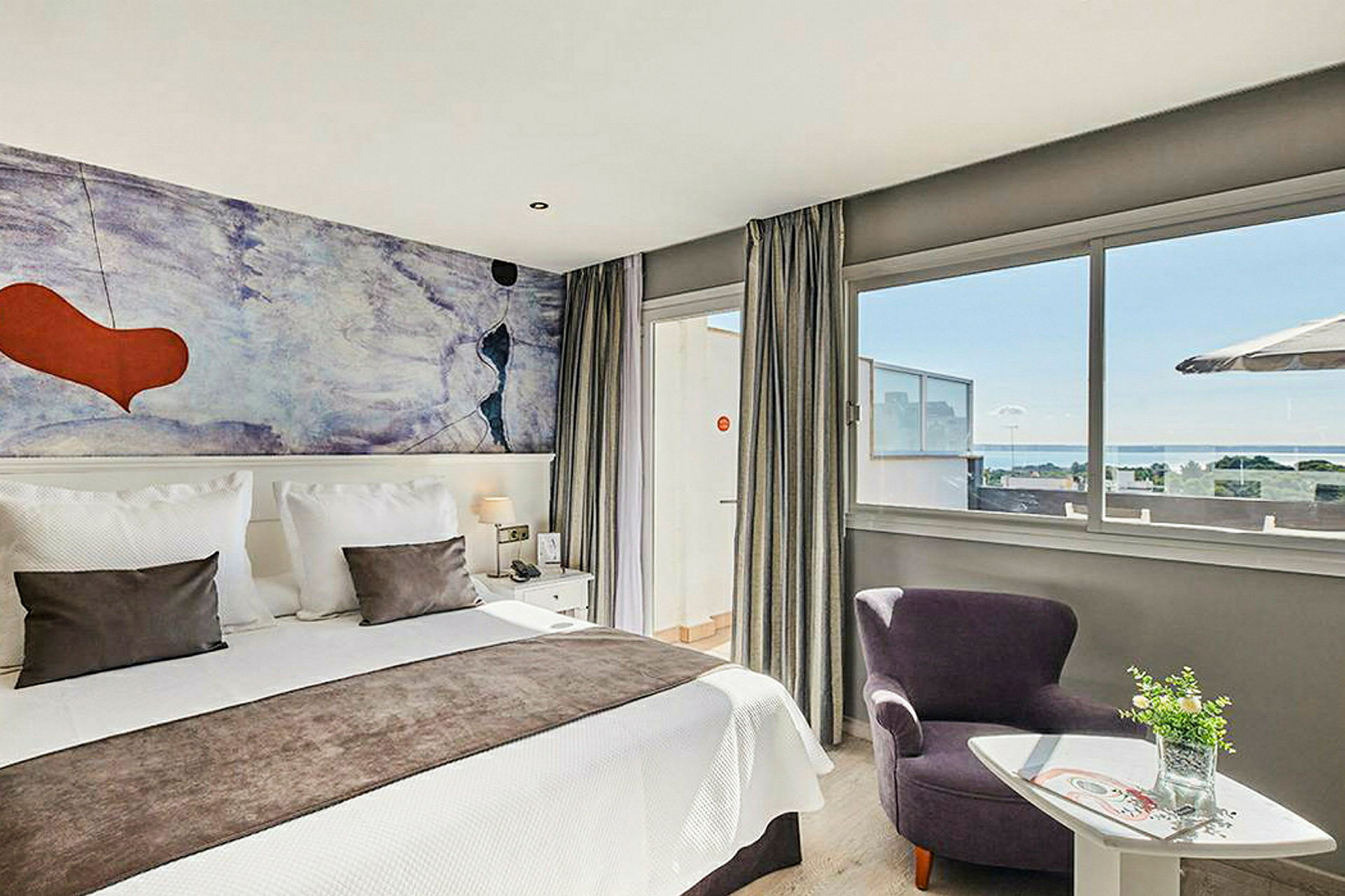 Et moderne hotelværelse med en dobbeltseng, farverigt vægmaleri og store vinduer med udsigt over havet. (AI-genereret tekst)