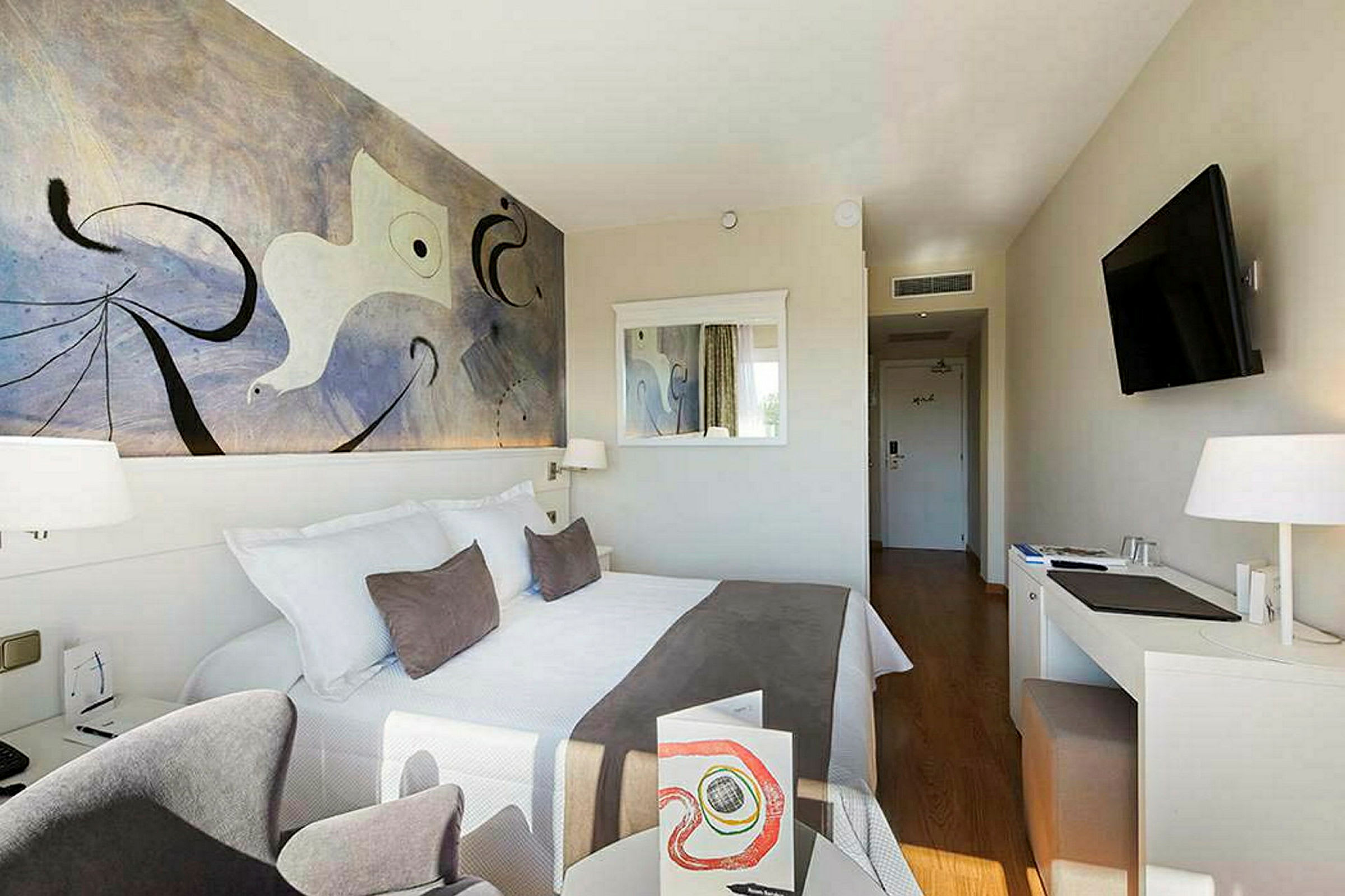 Et hotelværelse med en dobbeltseng, vægkunst, spejl, skrivebord og en TV. (AI-genereret tekst)