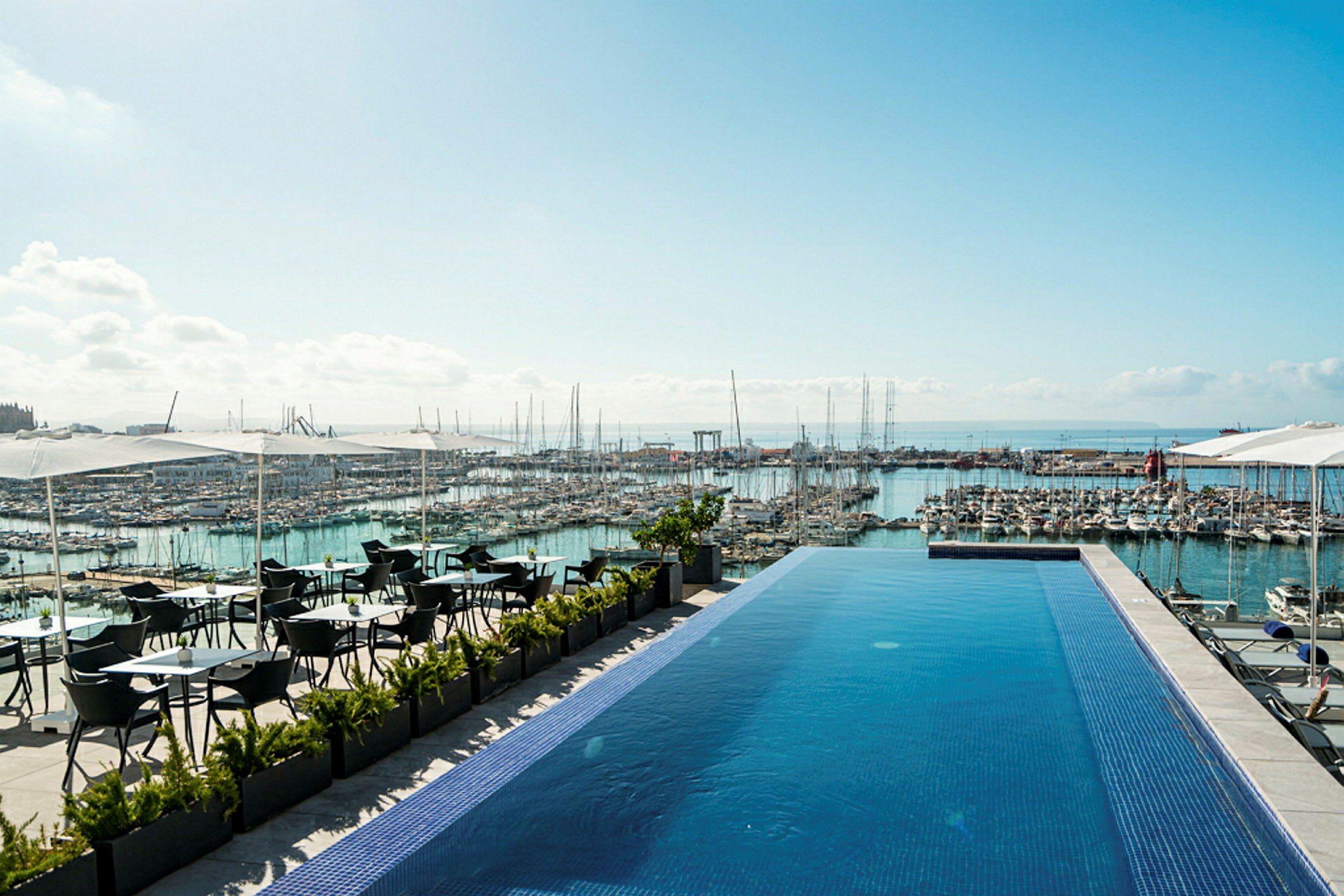 En infinitypool på en terrasse med udsigt over en marina fyldt med både og havet i baggrunden. Der er siddepladser under parasoller ved siden af poolen. (AI-genereret tekst)