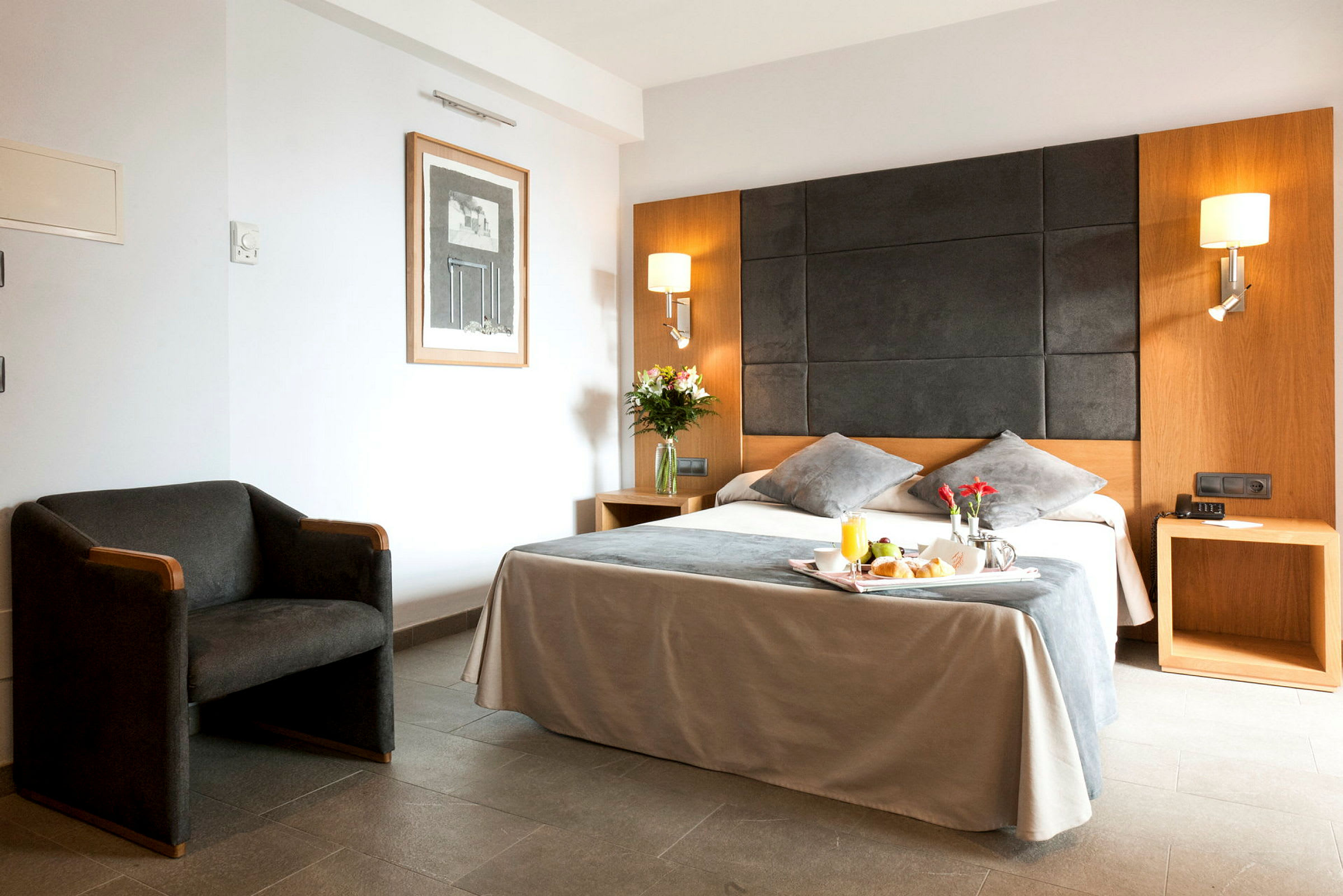 Et moderne hotelværelse med en stor seng, to natborde og en lænestol. På sengen er der en bakke med morgenmad. (AI-genereret tekst)