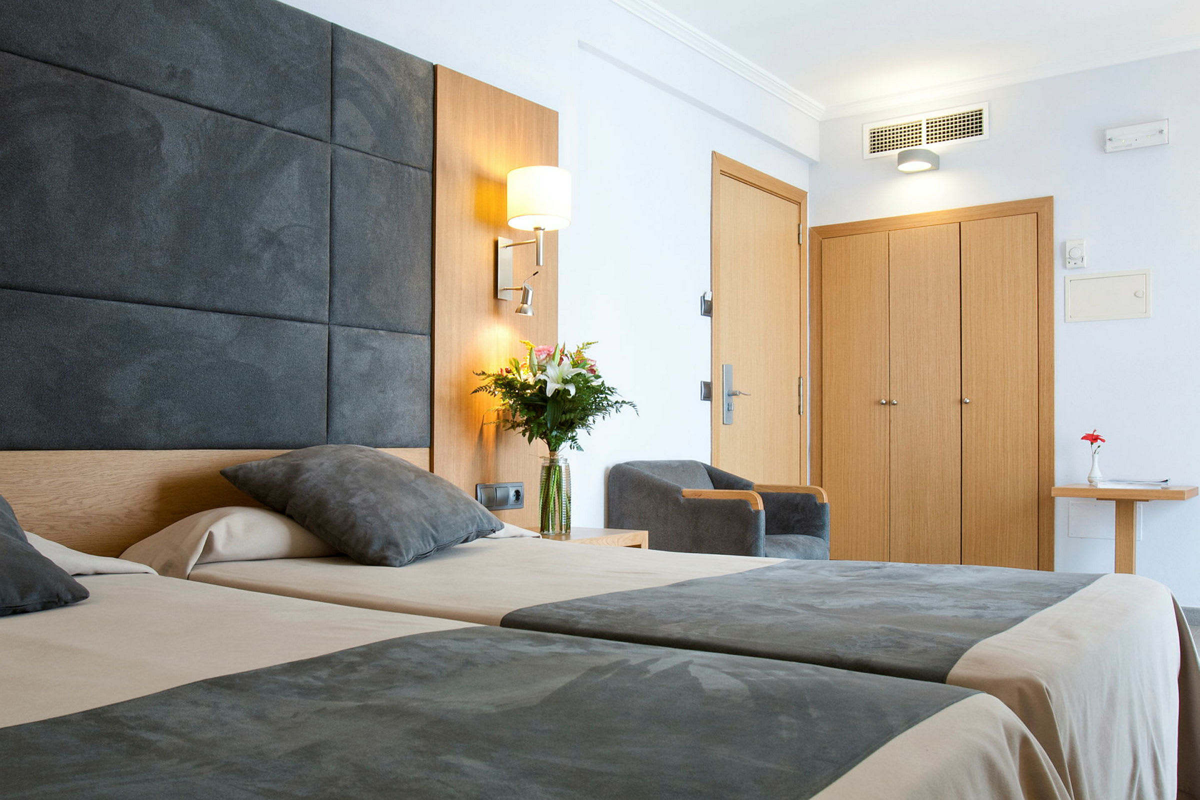 Billede af et moderne hotelværelse med to senge, grå sengetæpper, en stol, et træskab og en blomstervase på et bord. (AI-genereret tekst)